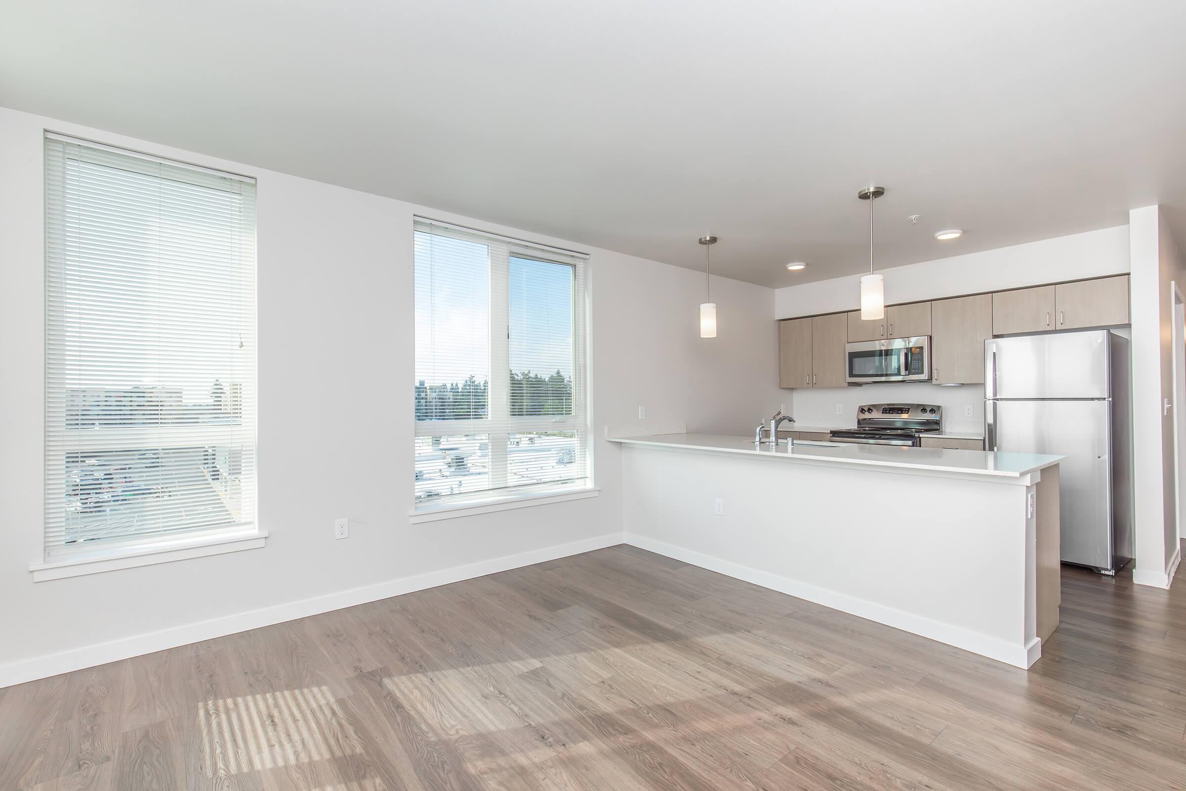 A modern kitchen and living area featuring large windows, light-colored walls, and hardwood flooring. The kitchen includes stainless steel appliances, a minimalist counter, and pendant lighting. The space is well-lit and airy, giving a spacious and contemporary feel.