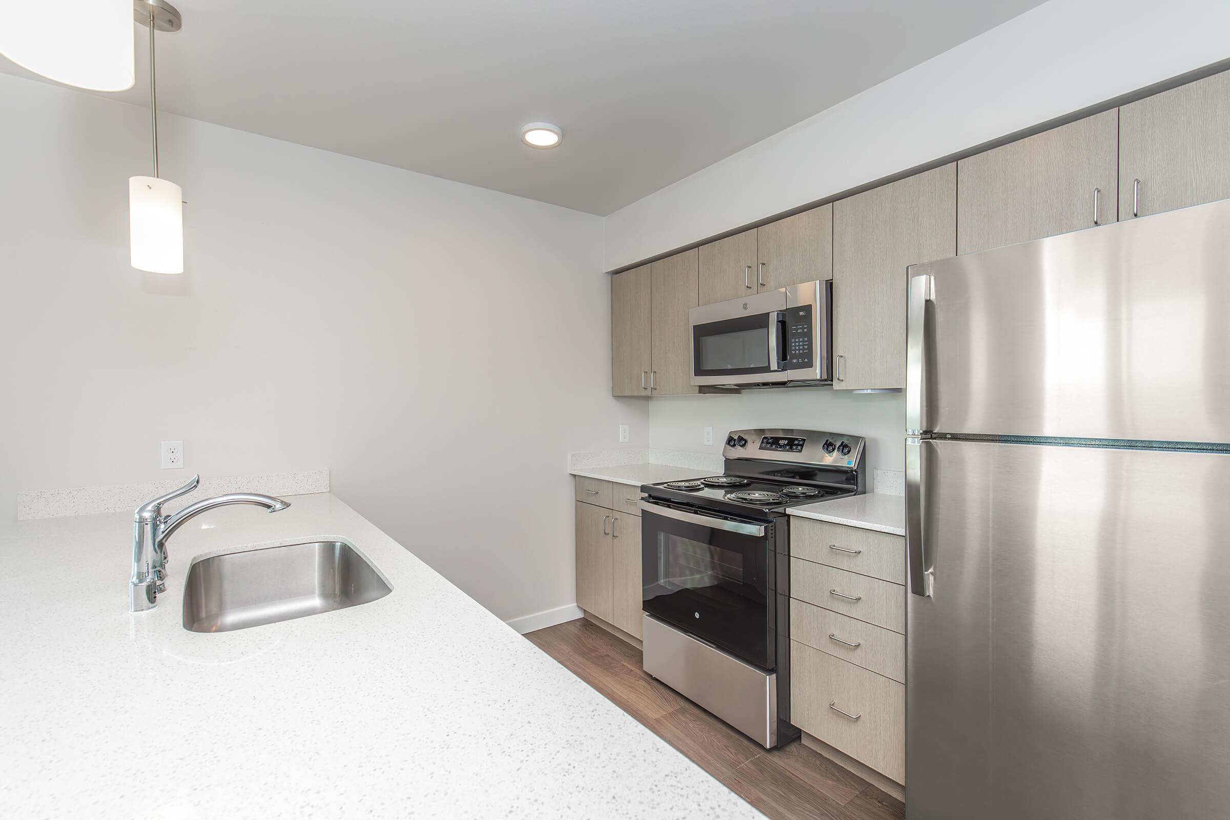Modern kitchen featuring light wood cabinetry, stainless steel appliances including a microwave and refrigerator, and a sleek countertop. The space is well-lit with a pendant light and includes a sink integrated into the countertop, creating a clean and contemporary look.