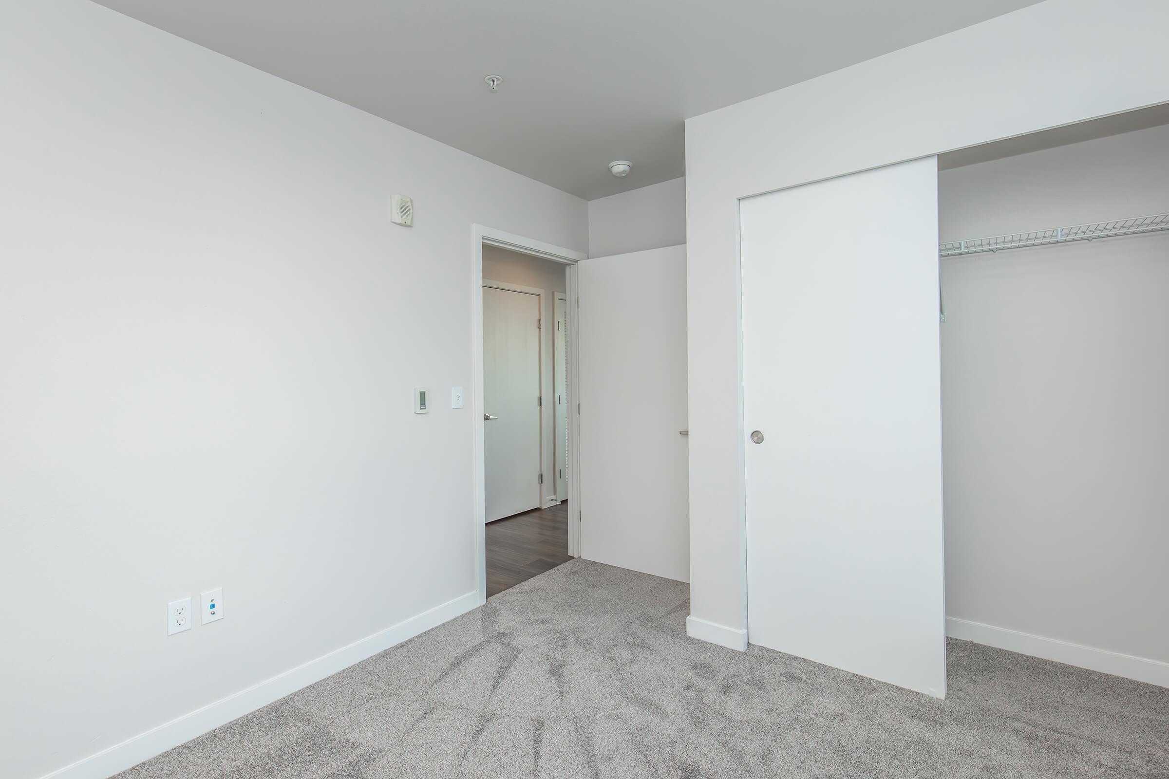 A clean, empty room featuring light gray walls and carpet flooring. There are two doorways: one leading to another room and another to a closet. The room has a modern, minimalist feel with bright lighting and no furniture or decorations.