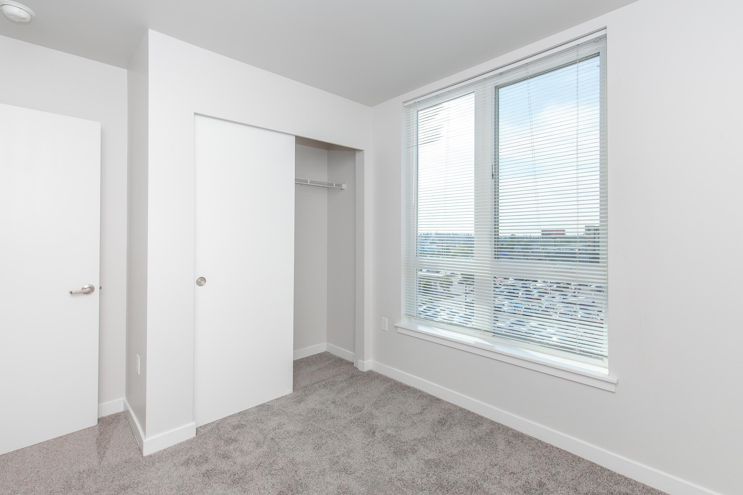 A modern, empty bedroom featuring light gray carpet and white walls. It includes a large window with blinds allowing natural light, a closet with sliding doors, and a simple door leading to another area. The space is bright and minimalist, suitable for various decor styles.