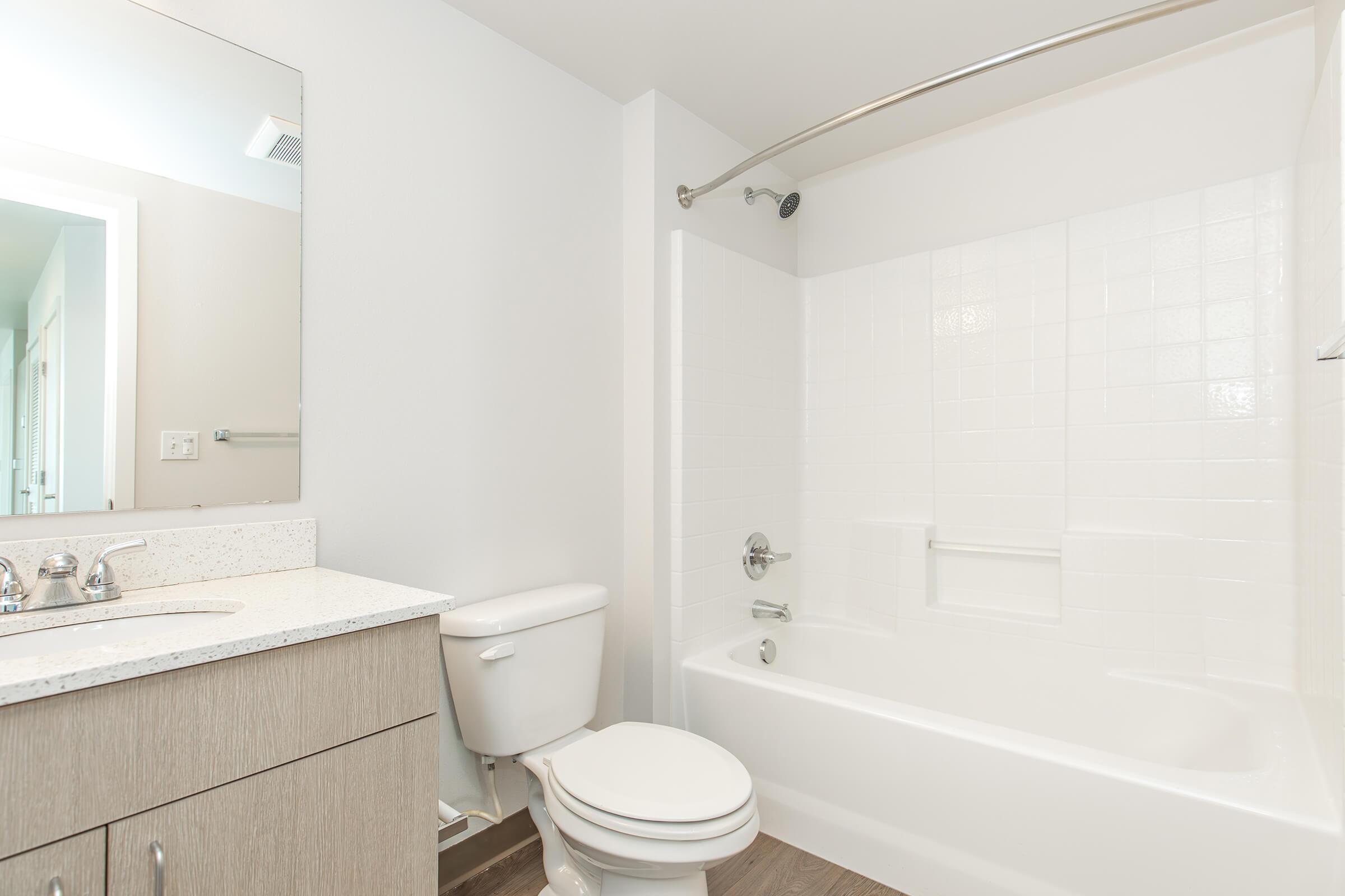 A clean, modern bathroom featuring a bathtub with a curved shower rod, a white tiled wall, a toilet, and a sink with a granite countertop. There is a large mirror above the sink and neutral-colored walls and flooring, creating a bright and spacious atmosphere.