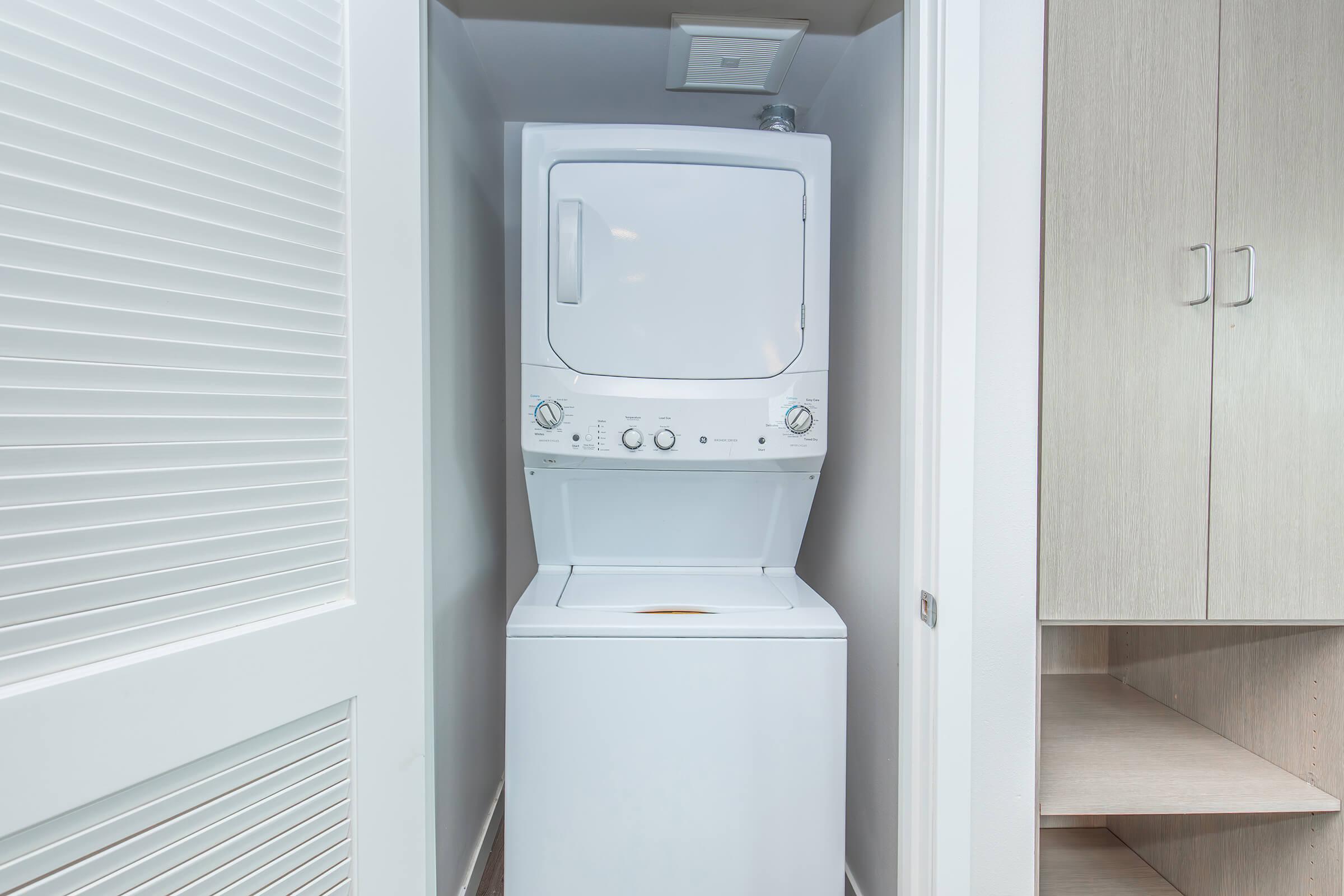 A stacked washer and dryer unit placed in a narrow laundry space, with white cabinetry and a door partially visible on the left side. The appliances have control knobs and display panels clearly visible. The overall setting is clean and modern, showcasing a typical home laundry area.