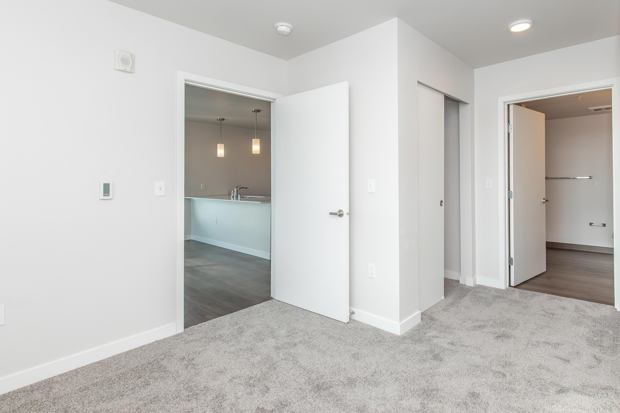 A modern interior space featuring a light gray carpet, white walls, and multiple doors leading to adjacent rooms. Two doors are open, revealing a kitchen area with pendant lighting in the background. The room has a bright and airy feel, with natural light illuminating the space.