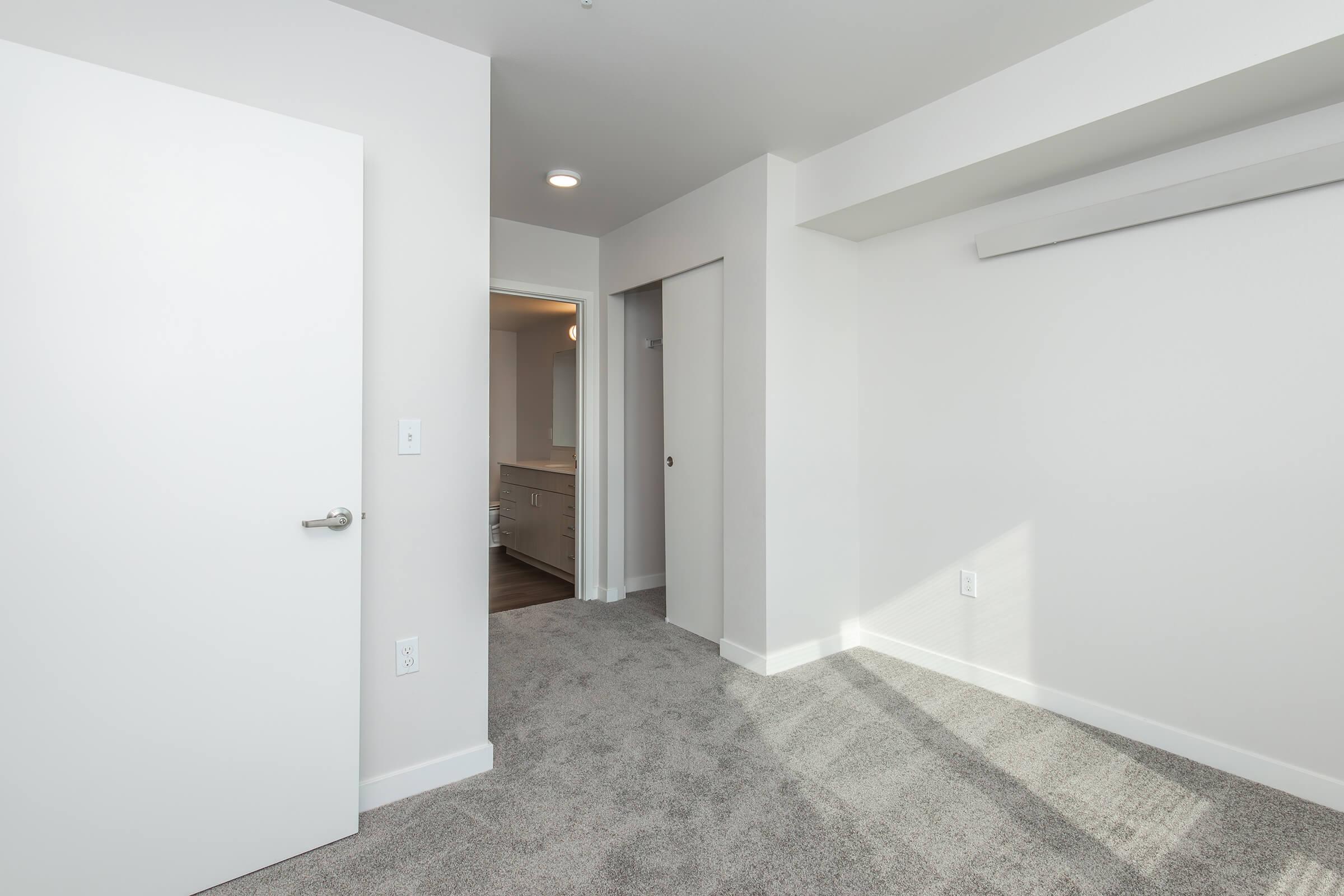 A modern, empty room with light gray walls and carpet. There is a closed white door on the left, leading outside, and a doorway at the back that opens to another room, possibly a bathroom. Natural light filters in from a window, highlighting the clean, minimalist design.