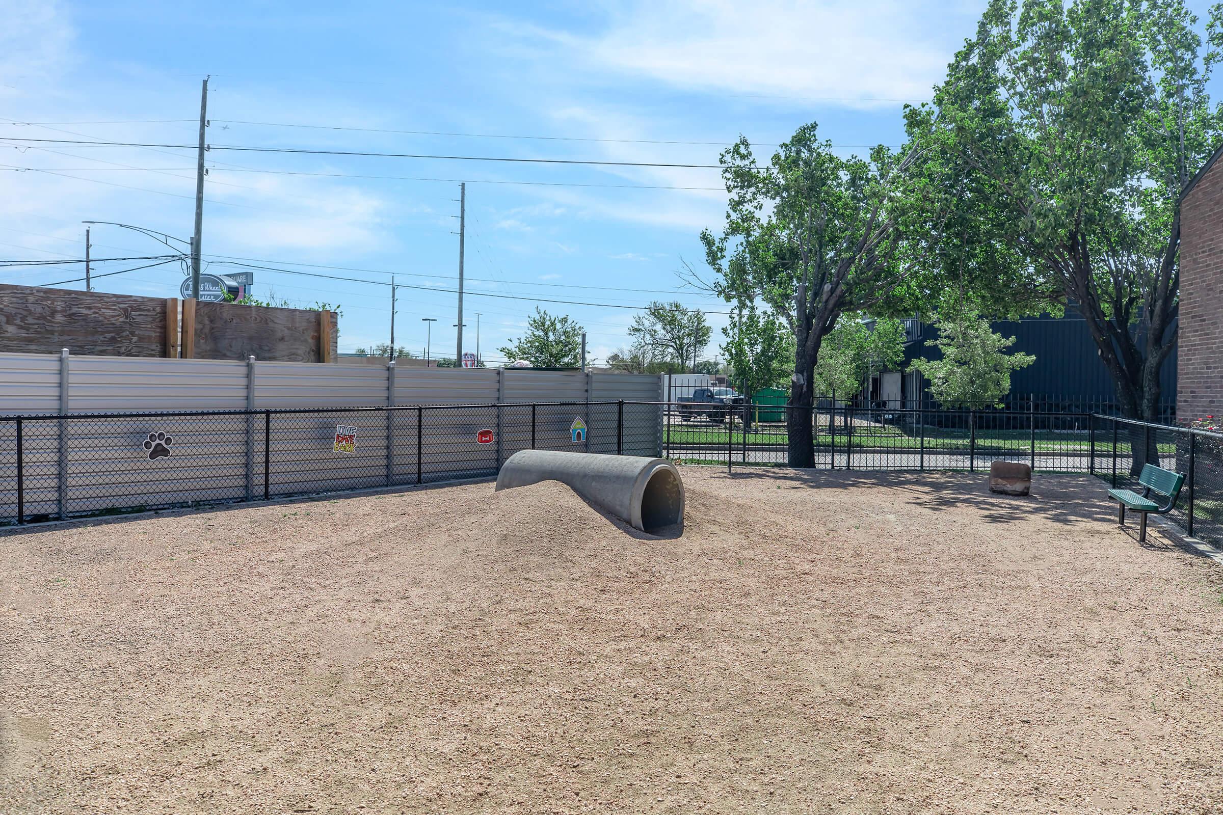 A sunny dog park featuring a sandy area with a large tunnel structure, surrounded by a black fence. Trees are in the background, providing shade, and there are a few benches for people to sit. Signs with paw prints are visible on the fence, indicating designated areas for pets.