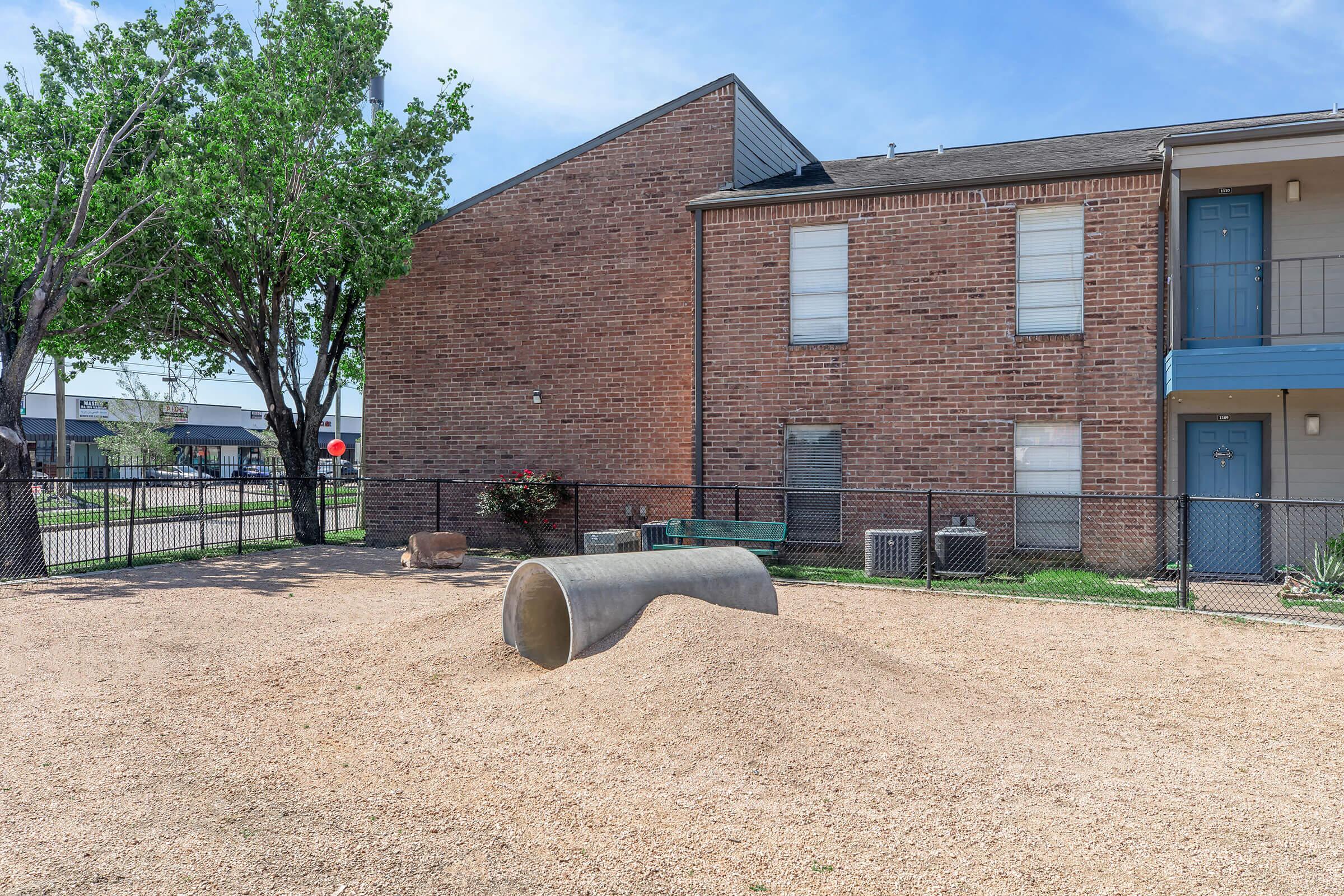 A playground area featuring a large concrete tunnel on a mound of gravel, surrounded by a fence. In the background, there are brick apartment buildings and trees, with blue skies overhead.