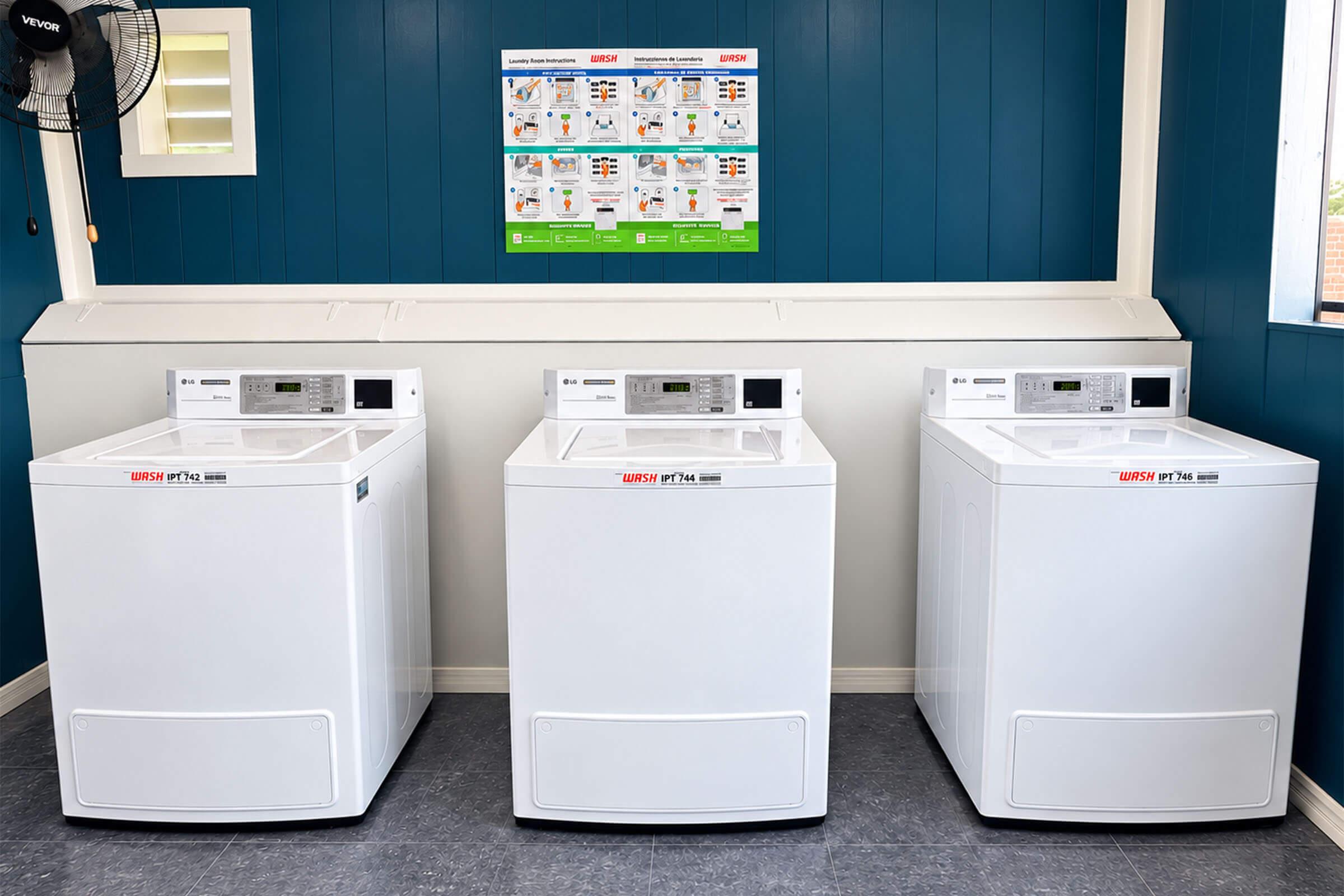 Three white washing machines arranged side by side in a laundry room. The walls are painted blue, and a poster with laundry instructions is hanging above the machines. The floor is a light gray material, and a ceiling fan can be seen in the corner of the room.