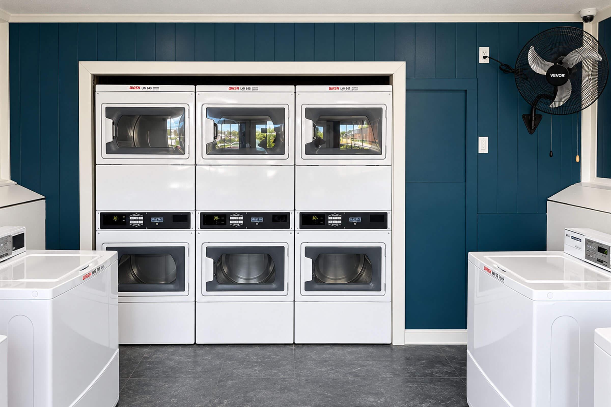 A modern laundry room featuring stacked washer and dryer units against a dark blue wall. The space includes white cabinetry and counter space, with additional washing machines placed nearby. Bright, well-lit environment with a ceiling fan for ventilation.