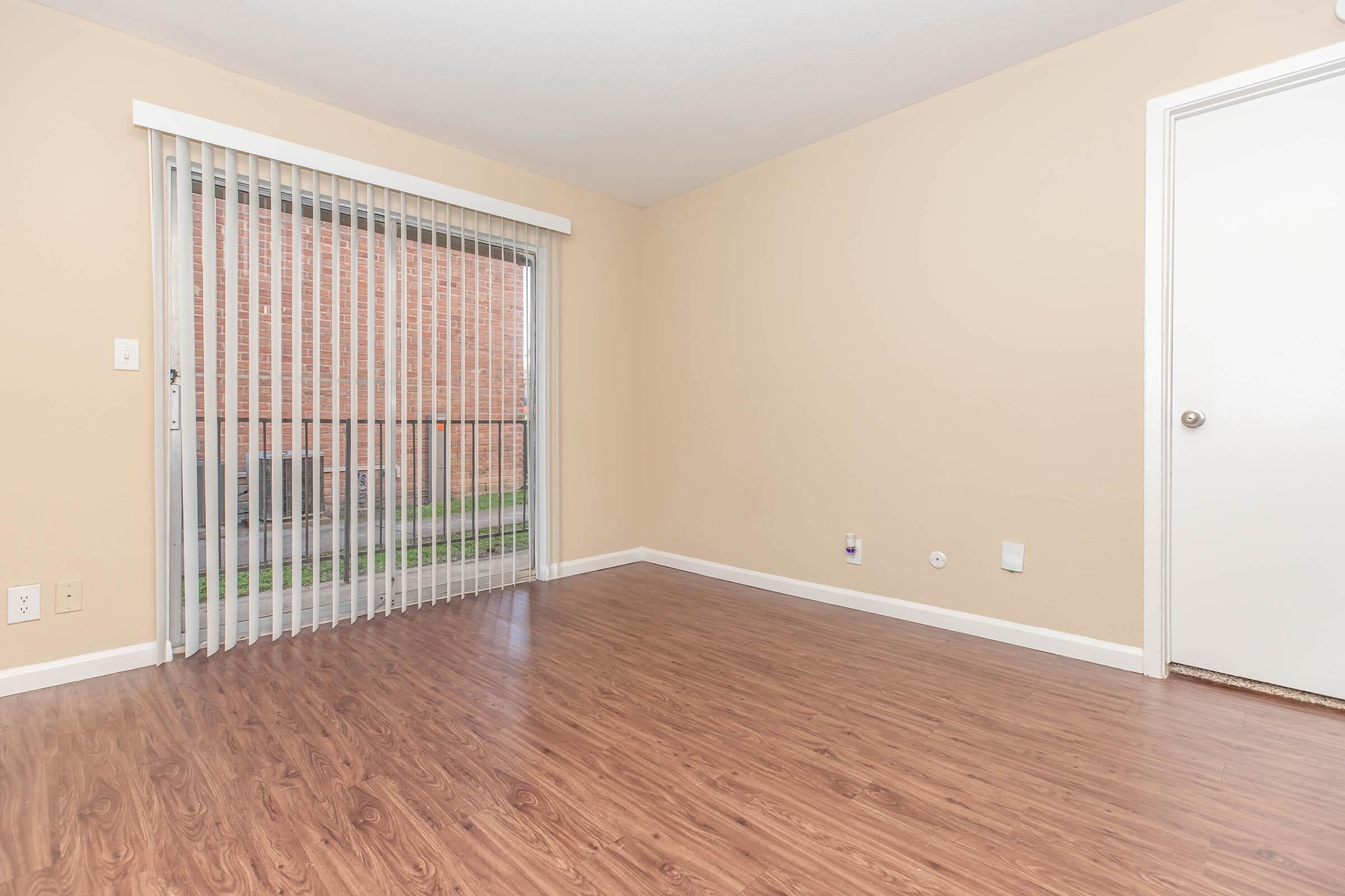 A spacious, empty room with light beige walls and a wooden floor. There are vertical blinds covering a large window that looks out onto a brick wall. A closed white door is visible on the right side of the image. The room is well-lit, creating an open and airy atmosphere.