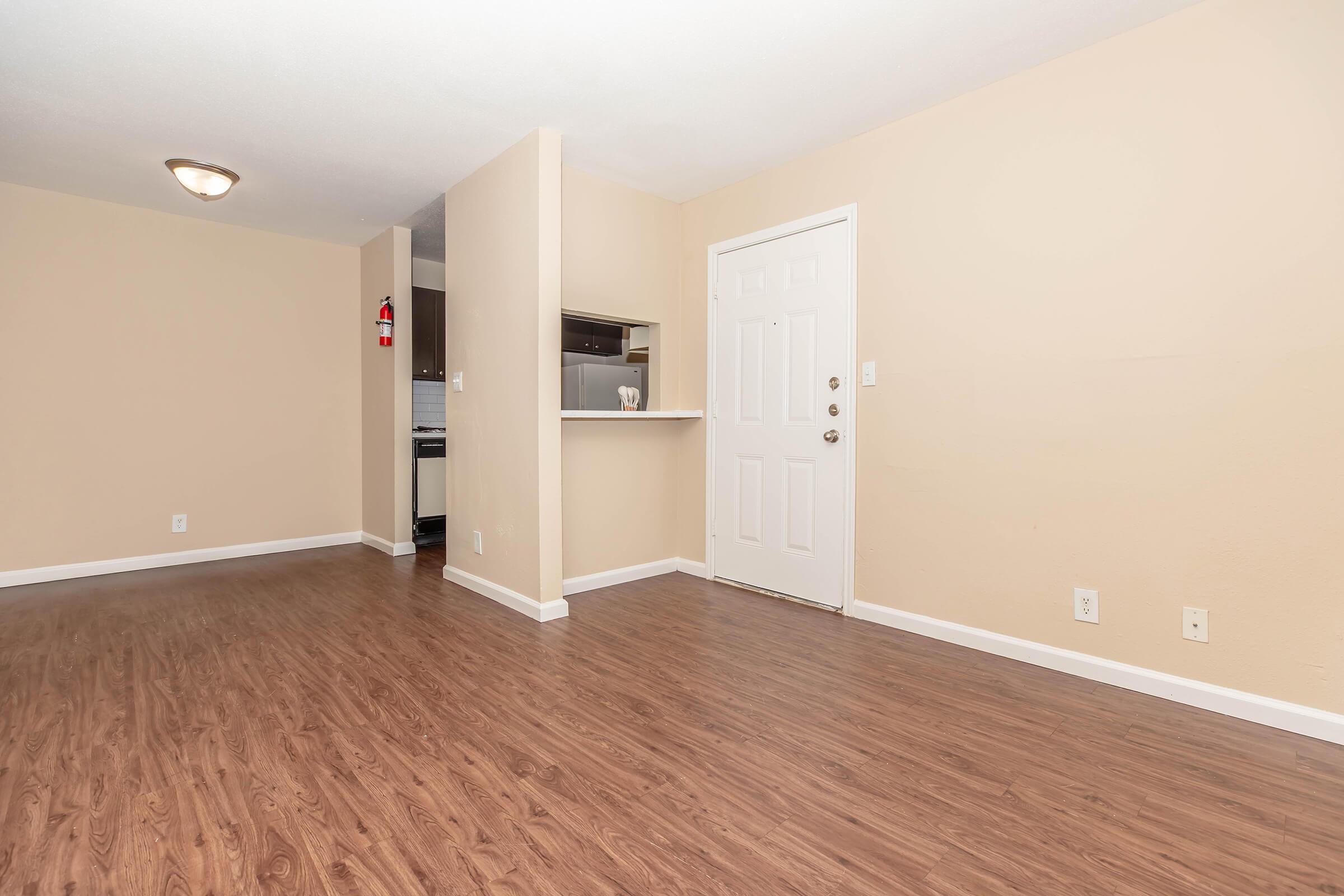 A light-colored room with laminate flooring, featuring a doorway leading to a kitchen area. One wall is painted beige, while another has a door and a simple light fixture. The space appears empty and well-lit, with a welcoming atmosphere.