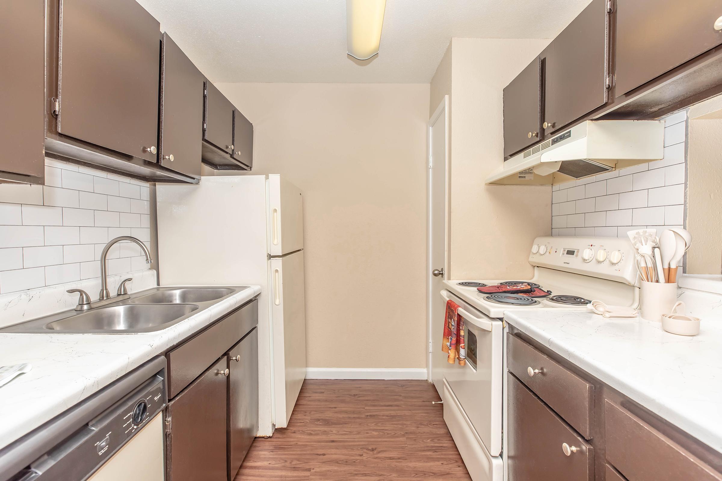 A modern kitchen featuring dark wood cabinets, a white countertop, and stainless steel appliances. The space includes a double sink, a refrigerator, an oven with a stovetop, and ample preparation area. Neutral-colored walls enhance the inviting atmosphere, and the floor is elegantly finished in a dark wood laminate.