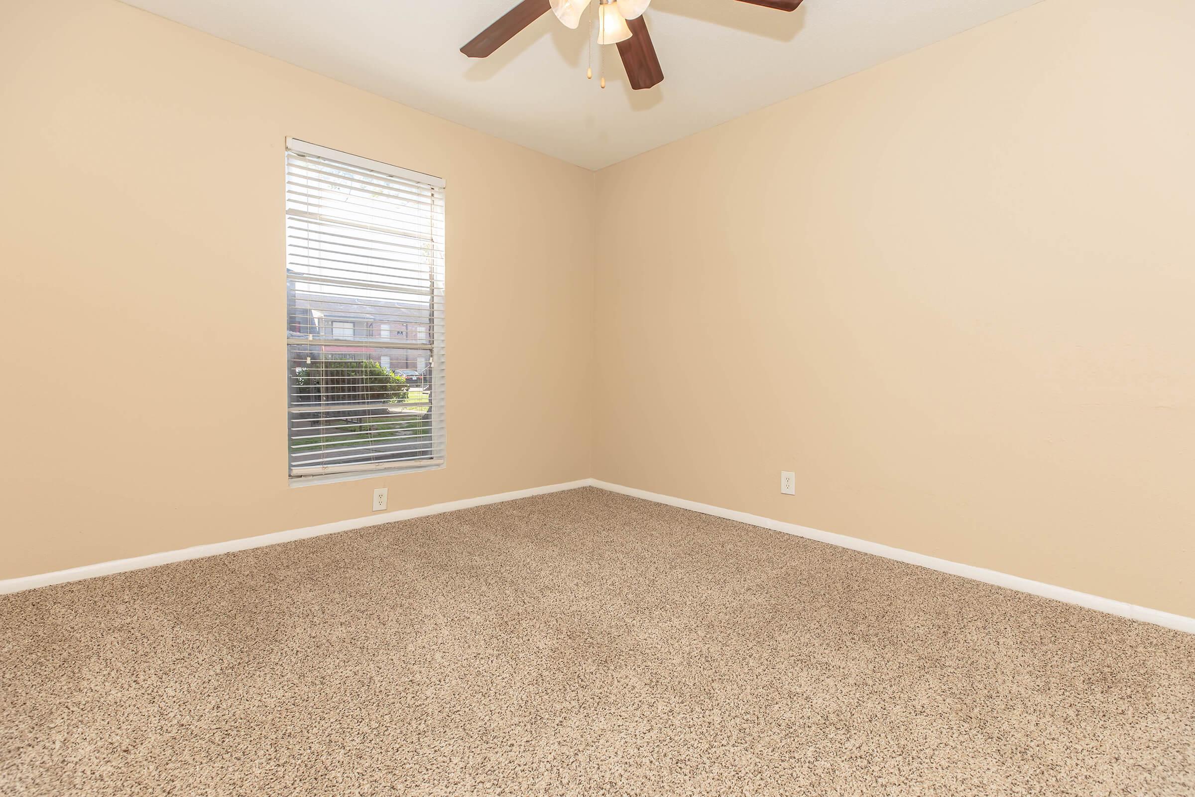 A vacant room with light beige walls and tan carpet. A ceiling fan is installed, and there is one window with white blinds that lets in natural light. The space appears clean and empty, suitable for decoration or furnishing.