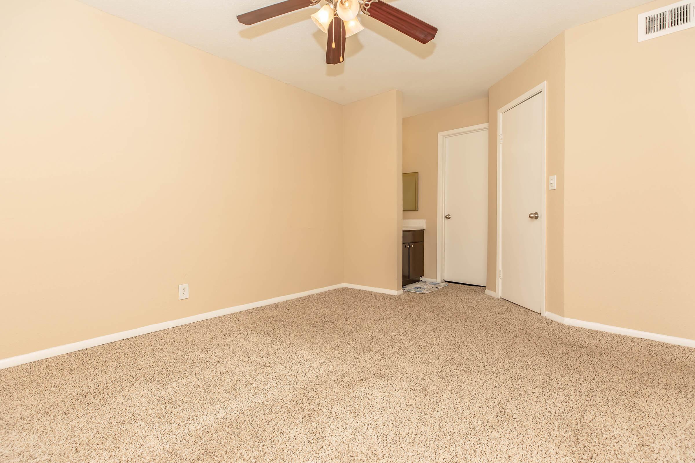 An empty room with light beige walls and carpet. A ceiling fan is mounted in the center. A small area with a sink and mirror is visible on the left, and a closed door is on the right leading to another area. The room appears well-lit and spacious, with no furniture present.