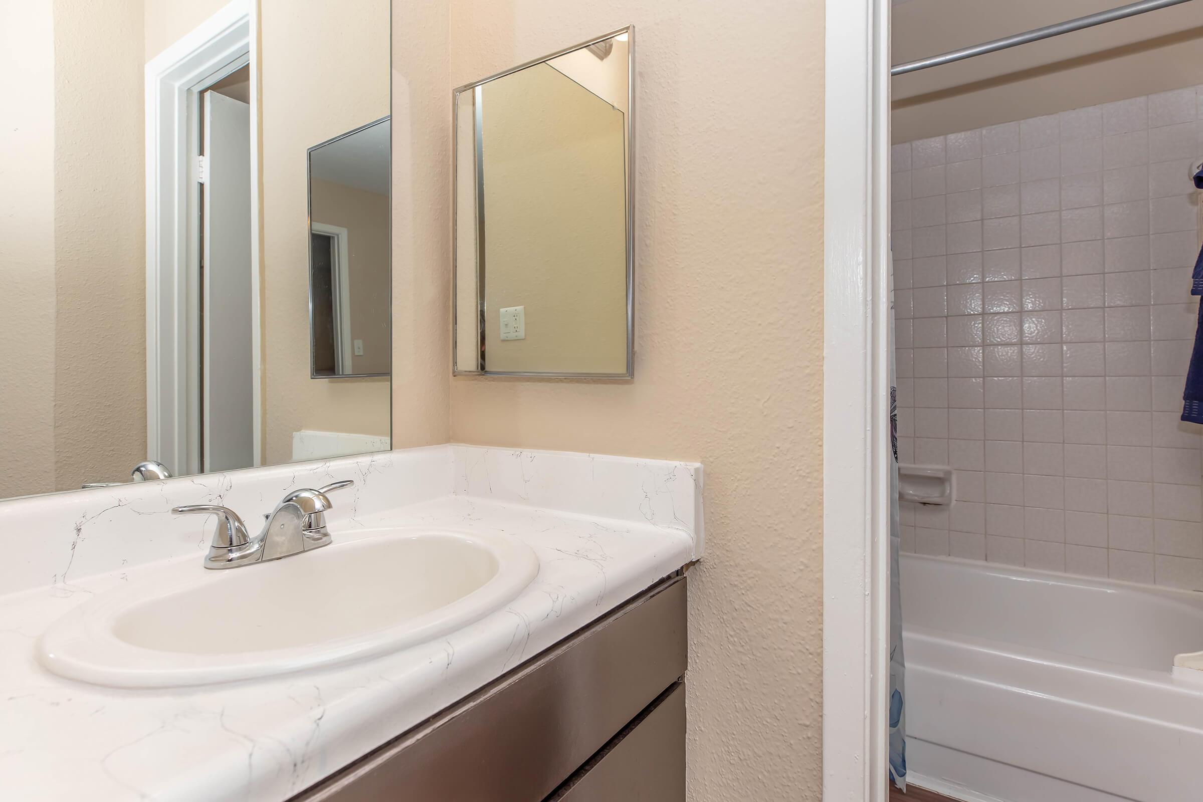 A clean bathroom featuring a white sink with a chrome faucet, a mirror above the sink, and a light-colored countertop. The adjacent space includes a shower/tub combination with a tiled wall, and neutral-colored walls that create a bright, airy atmosphere.