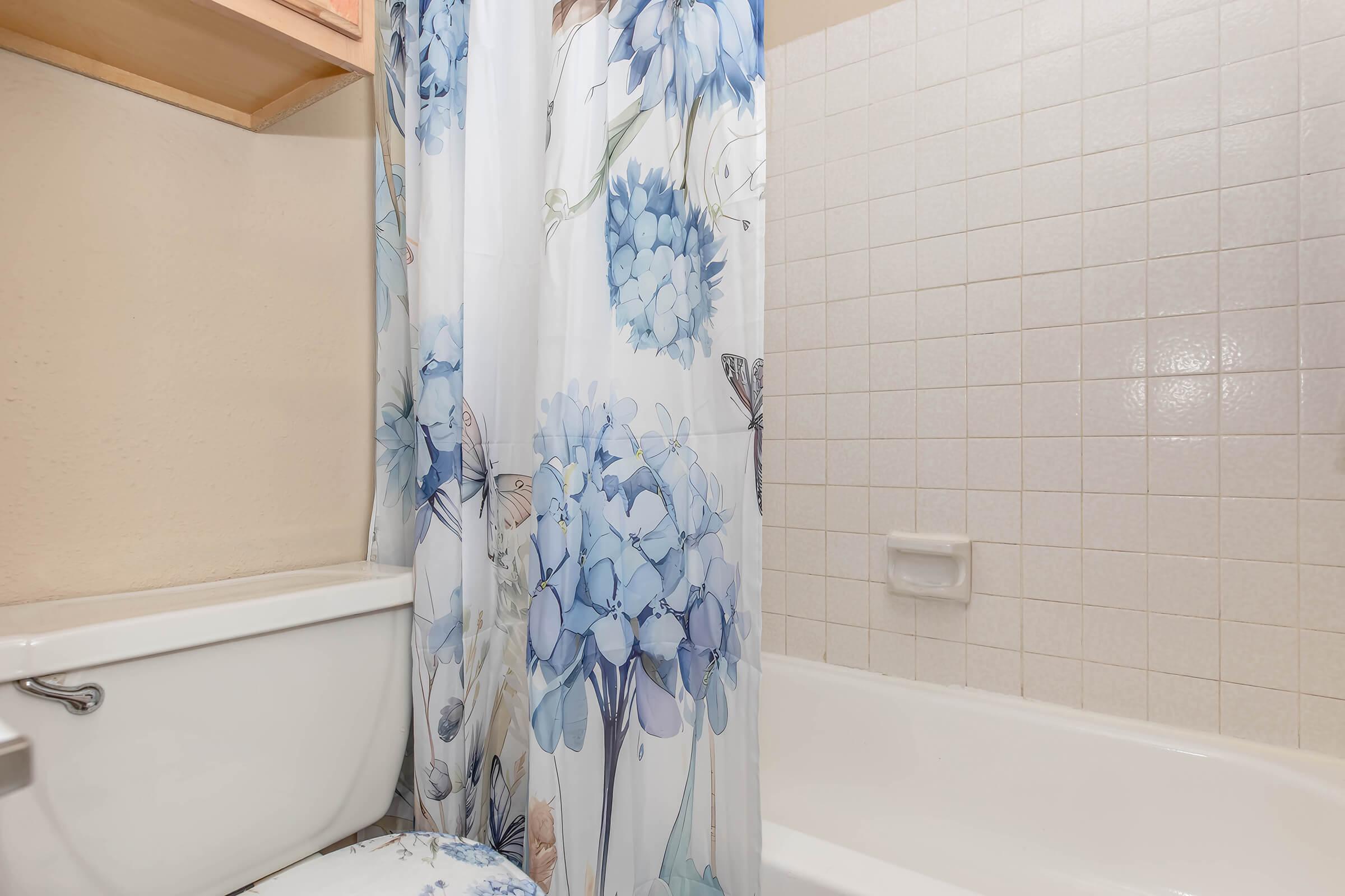 A bathroom featuring a white bathtub, a toilet, and a floral shower curtain with blue hydrangeas. The wall tiles are light-colored, and there is a small shelf above the toilet. The overall ambiance is clean and bright.