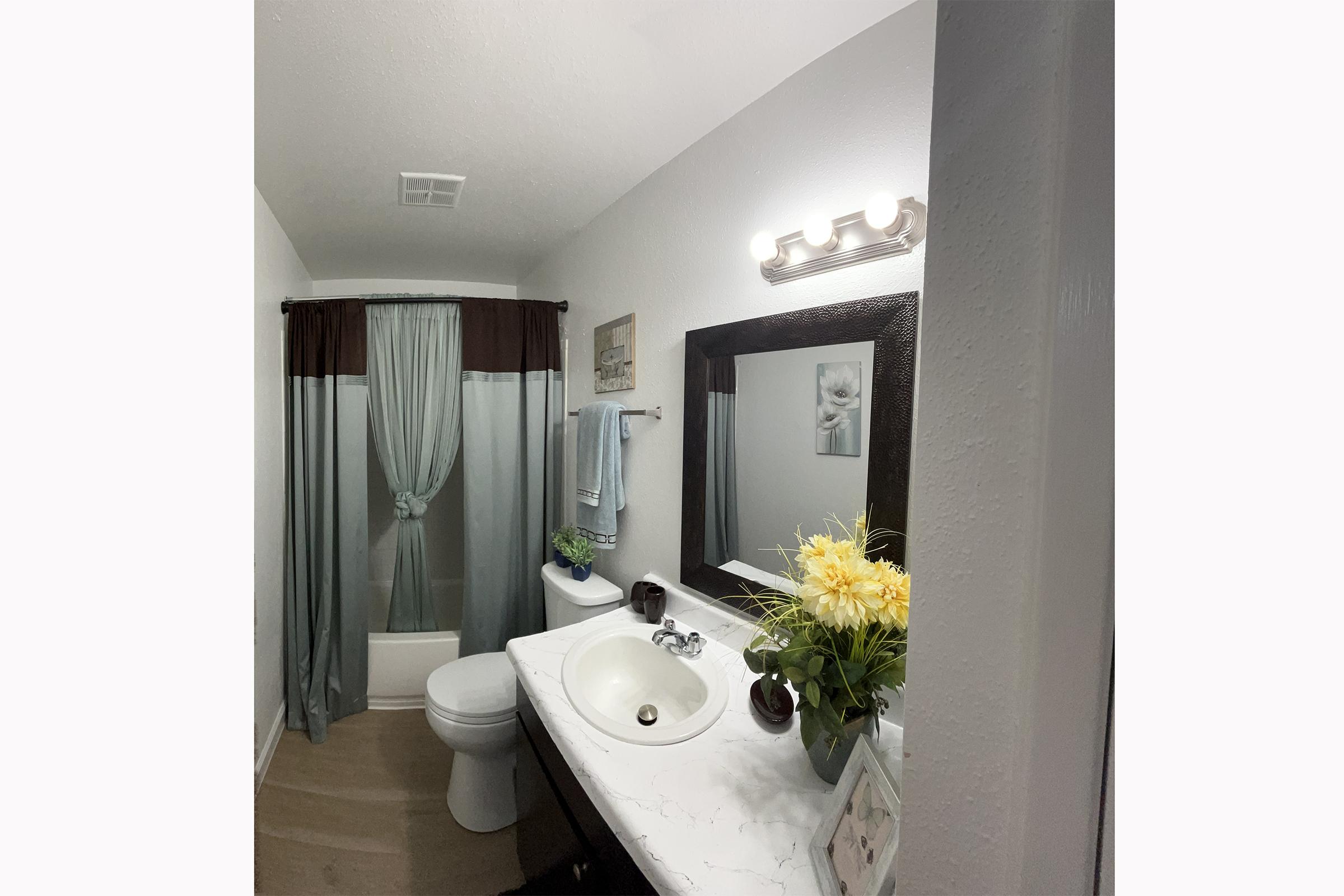 A clean bathroom featuring a marble countertop with a sink and faucet, a large mirror above the sink, a white toilet, and decorative elements. The space has gray curtains, a blue towel hanging on a towel rack, and a small bouquet of yellow flowers on the counter, enhancing the inviting atmosphere.