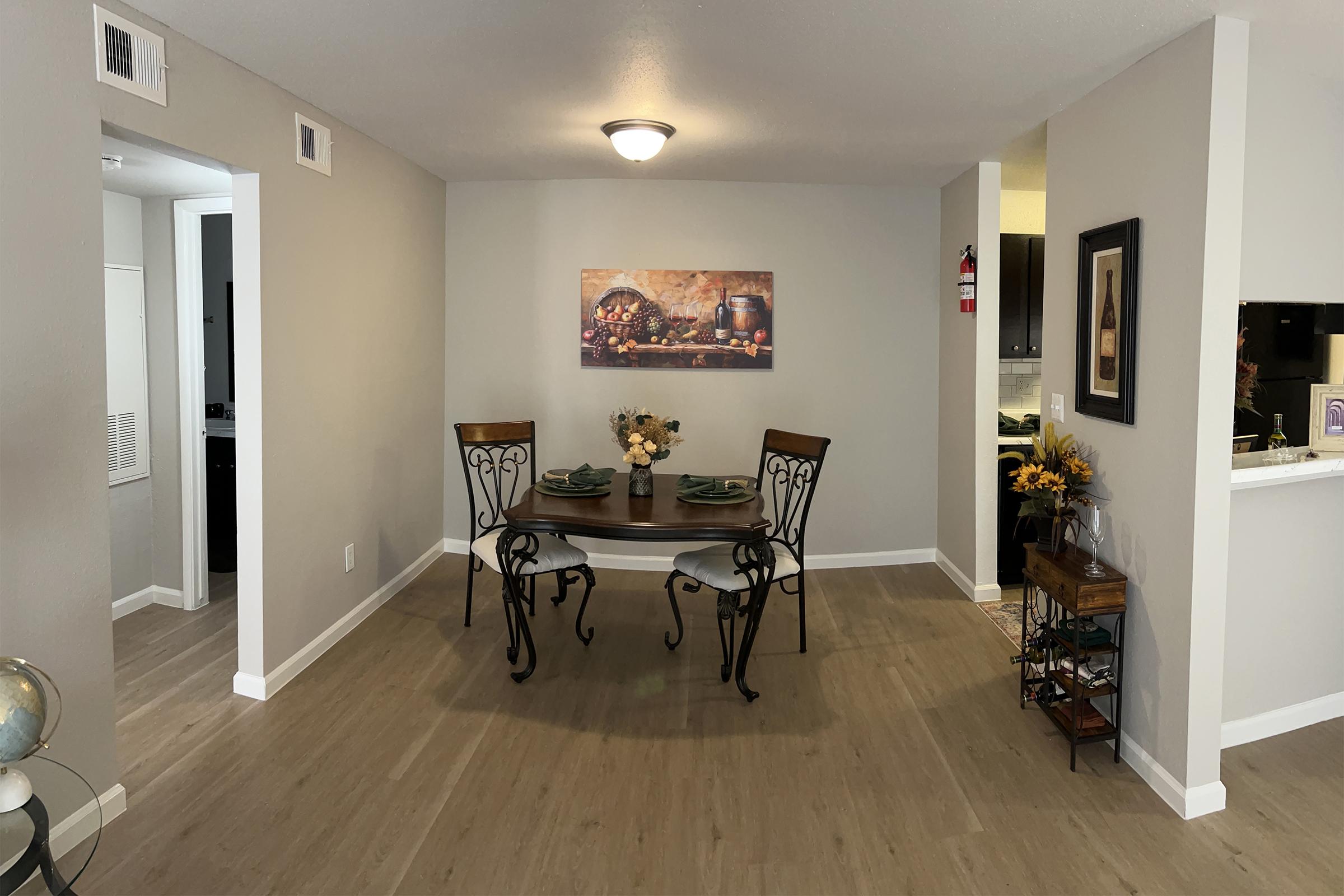 A cozy dining area featuring a small table set for two with elegant tableware. A floral centerpiece adorns the table, and a colorful artwork hangs on the wall behind it. The room has neutral-colored walls, modern lighting, and wooden flooring, creating a warm and inviting atmosphere. An adjacent kitchen is partially visible.