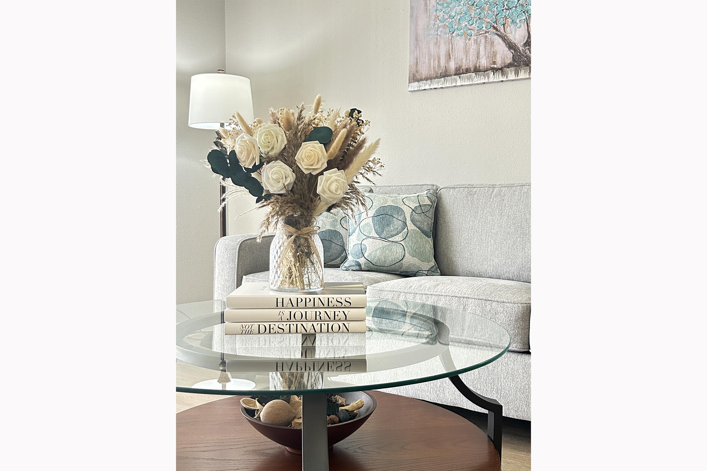 A modern living room scene featuring a glass coffee table with stacked books, a vase of white roses and dried flowers, and a cozy gray sofa with patterned cushions. A lamp and a piece of wall art depicting a tree add warmth to the space.