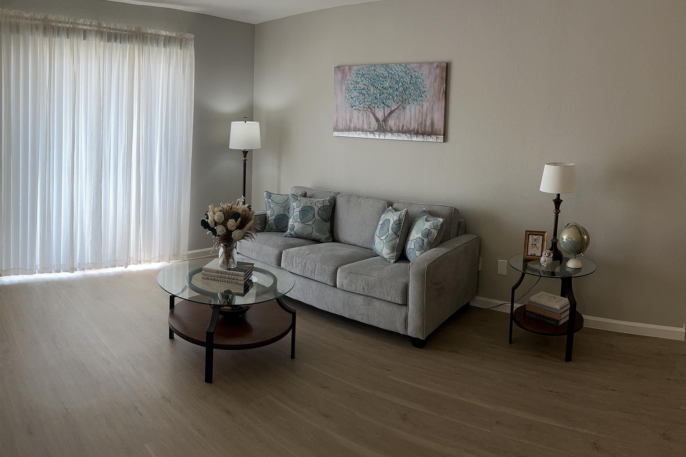 A cozy living room featuring a light gray sofa with decorative pillows, two side tables with lamps, a glass coffee table with a floral arrangement and a globe, and large windows with sheer curtains allowing natural light to fill the space.