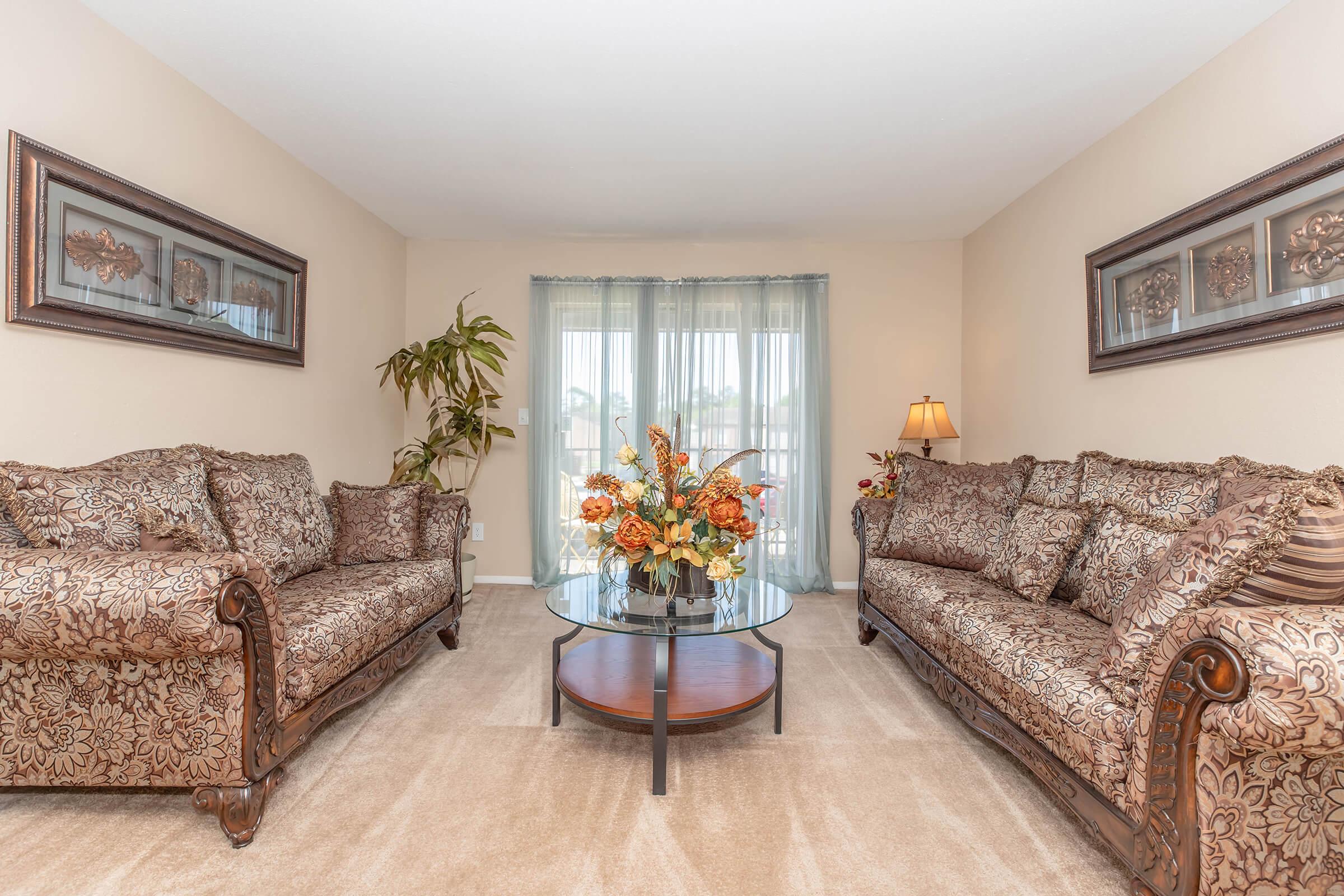 Cozy living room featuring two ornate brown sofas with floral patterns, a glass coffee table with a floral arrangement, and a soft beige carpet. Natural light filters through sheer curtains, creating a warm and inviting atmosphere, complemented by framed wall art and a decorative lamp.