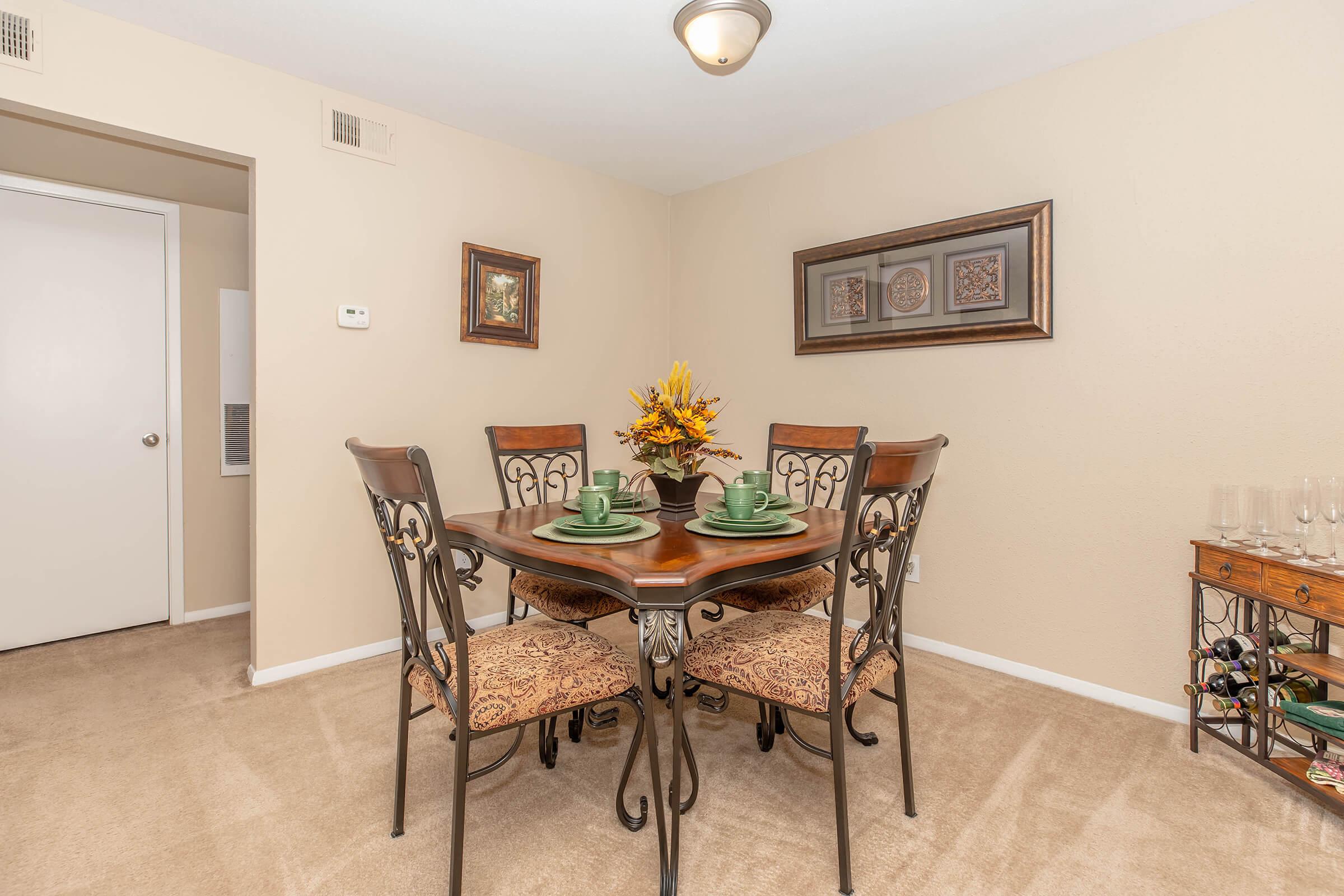 A cozy dining area featuring a wooden table surrounded by four ornate chairs, each with floral patterned upholstery. The table is set with green plates and a decorative centerpiece of sunflowers. The walls are painted in a neutral tone, and there are framed artworks and a wine rack in the background.