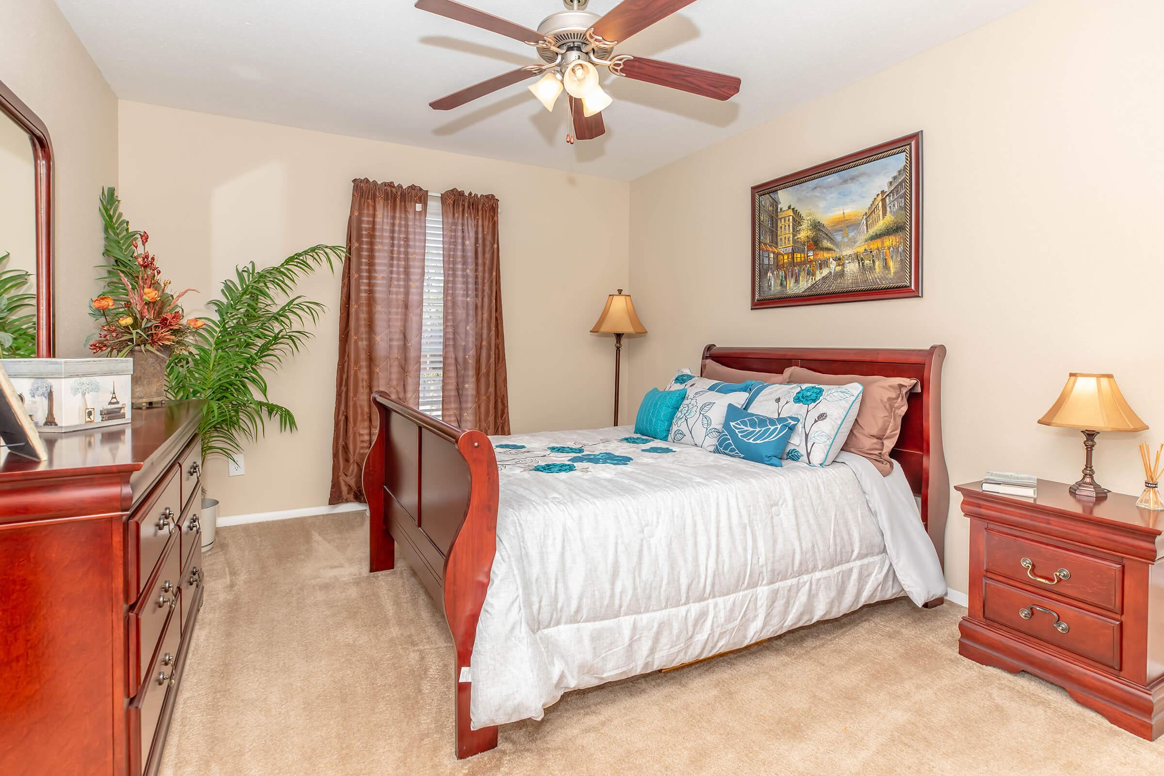 A cozy bedroom featuring a wooden bed with decorative blue pillows, a dresser, a bedside table with a lamp, and a mirror. The room is light-colored with beige walls and soft carpet. There are two windows with brown curtains and a potted plant, creating a warm and inviting atmosphere.