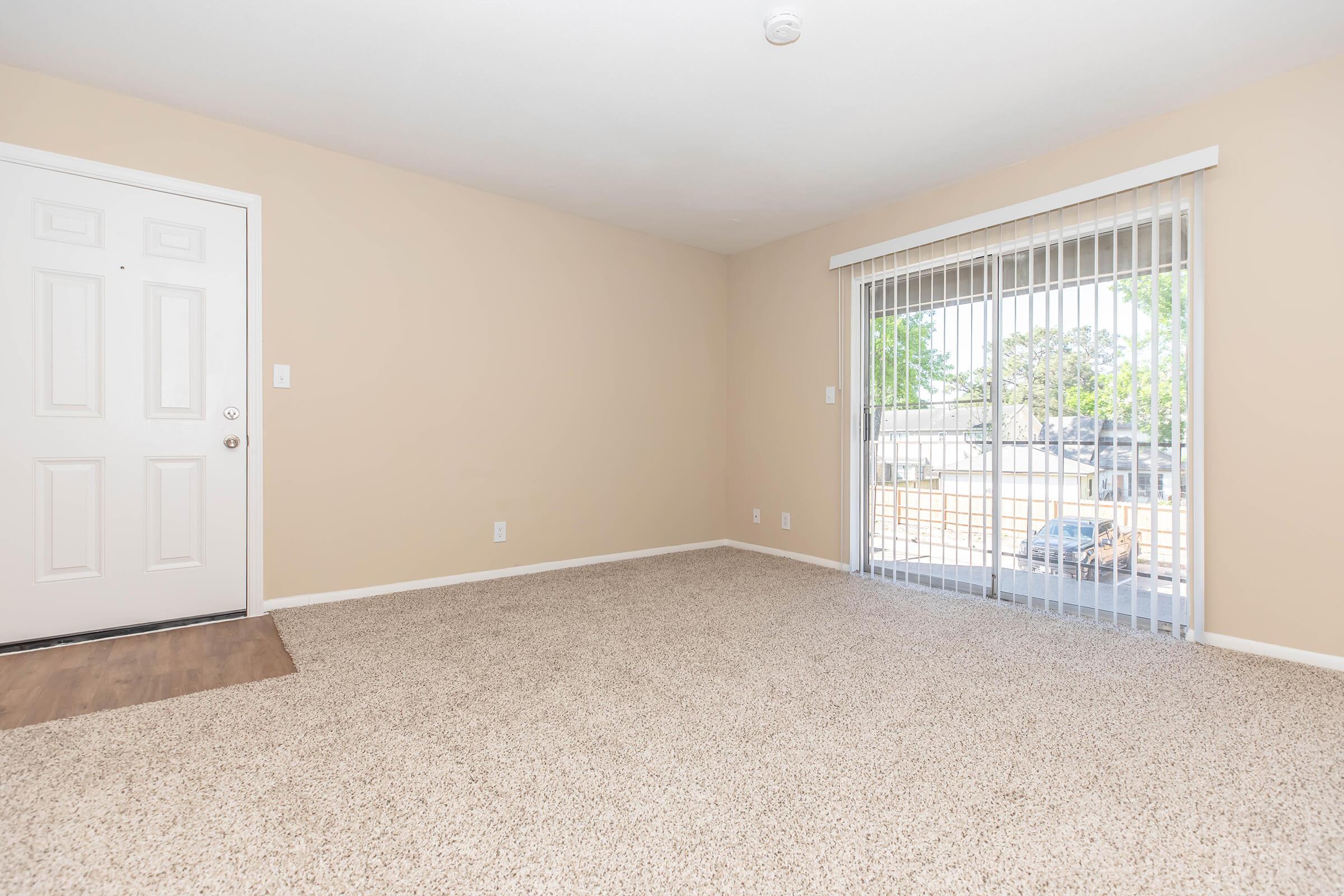 A spacious, empty living room with beige walls and light-colored carpet. On one side, there is a white front door, and on the opposite wall, large sliding glass doors lead to an outside area. Natural light filters in through the doors, enhancing the open and airy feel of the space.