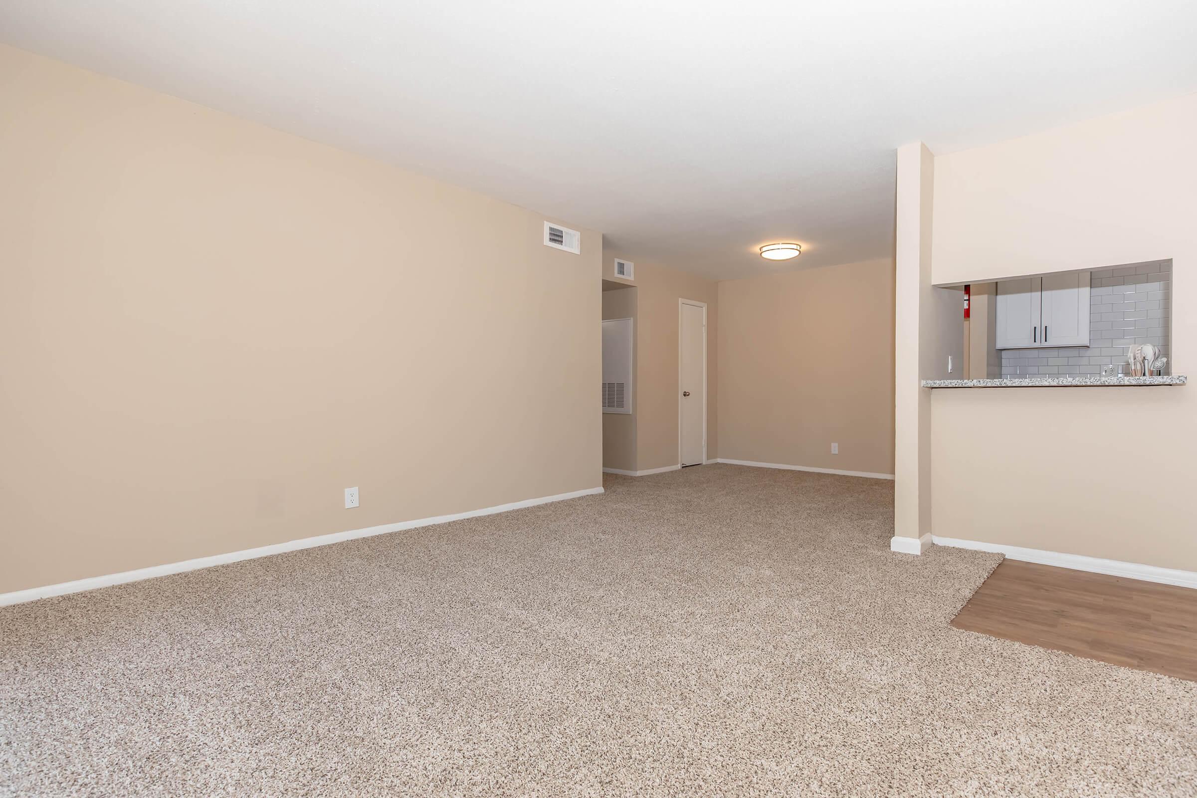 A spacious, empty living room with light beige walls and carpeting. There is a small kitchen area in the background with a bar countertop. The room has a single door leading to another space, and a ceiling light fixture is visible. Natural light may be coming in from an unseen window.