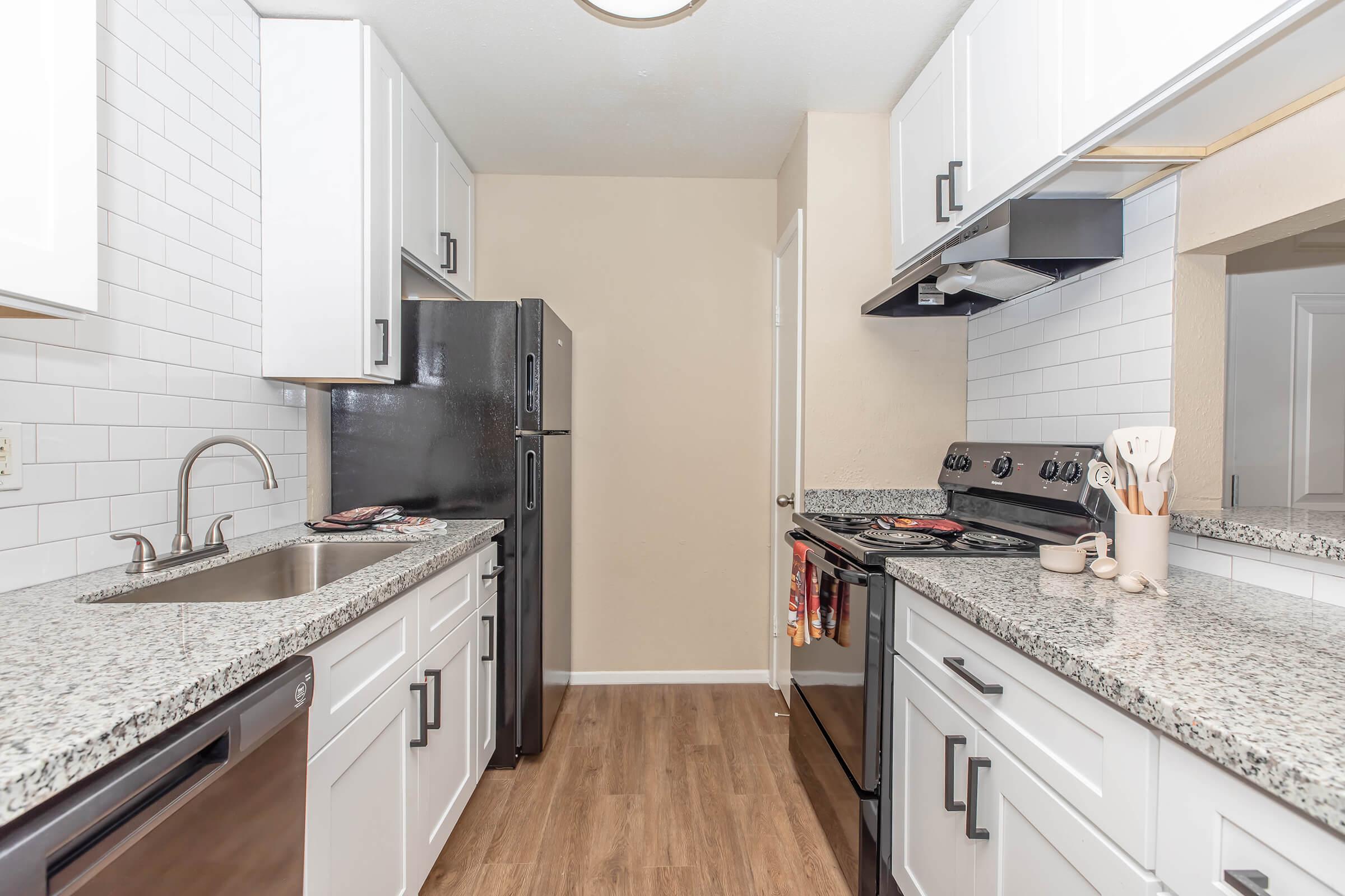 Modern kitchen with white cabinets, granite countertops, and stainless steel appliances. Features include a black refrigerator, a stove with an overhead microwave, and a sink with a stylish faucet. The flooring is wood-like, and the walls are painted a light beige.