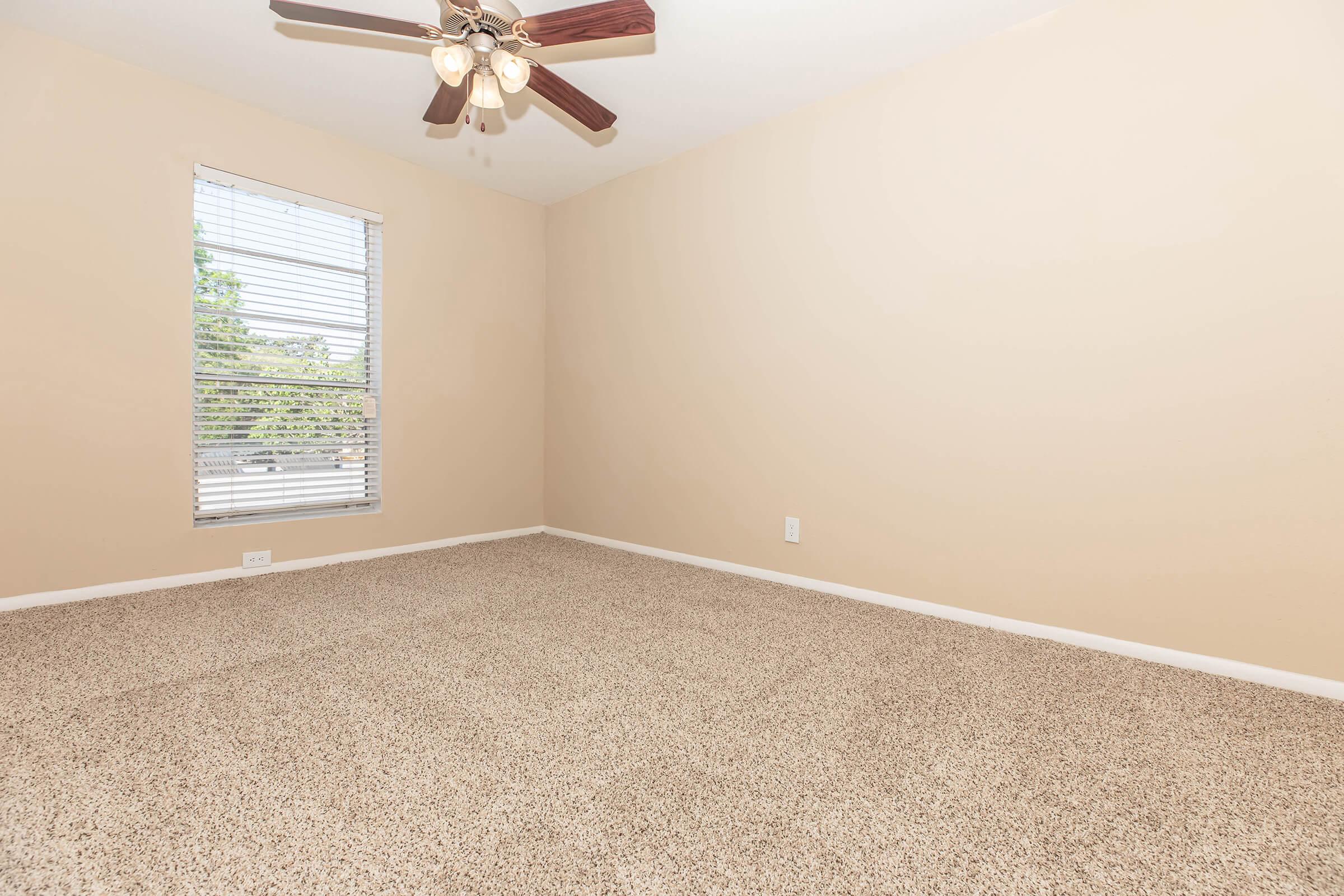 A spacious, empty room with light beige walls and a carpeted floor. A ceiling fan with wooden blades hangs from the ceiling. There is a single window with white blinds, allowing natural light to enter. The room has a clean and minimalistic appearance, ready for furniture and personal decoration.