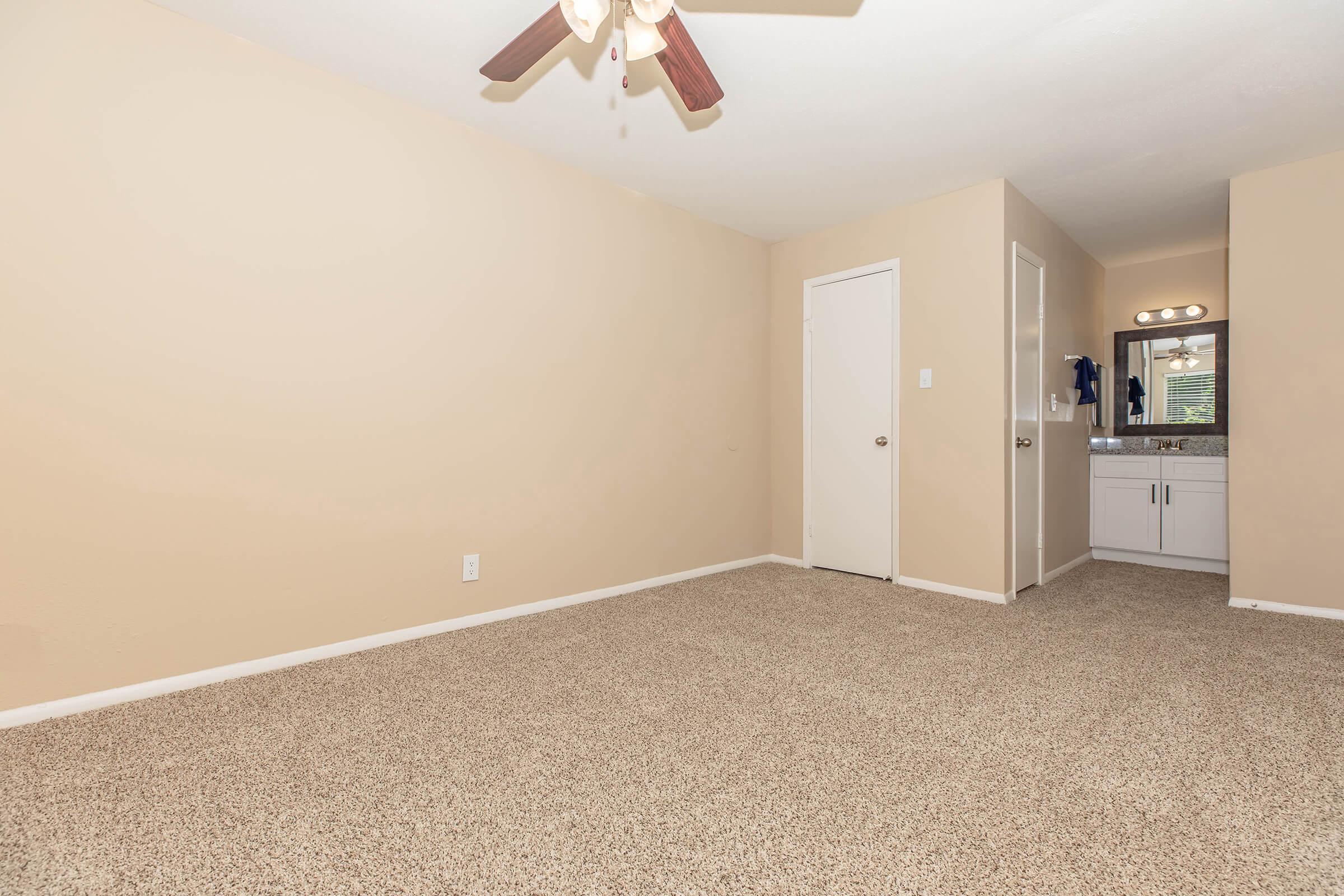 A spacious, empty room with beige walls and carpet. A ceiling fan is visible, and there's a doorway on the left leading to a bathroom area with a mirror and sink. Bright lighting enhances the open atmosphere, making it feel inviting and ready for new furnishings.