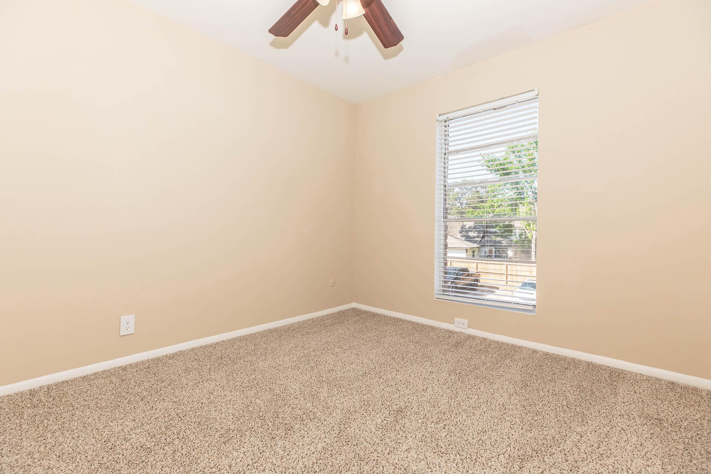 Empty room with beige walls, light brown carpet, and a ceiling fan. A window with blinds allows natural light to enter, offering a view of greenery outside. The space appears clean and well-lit, suitable for various uses.