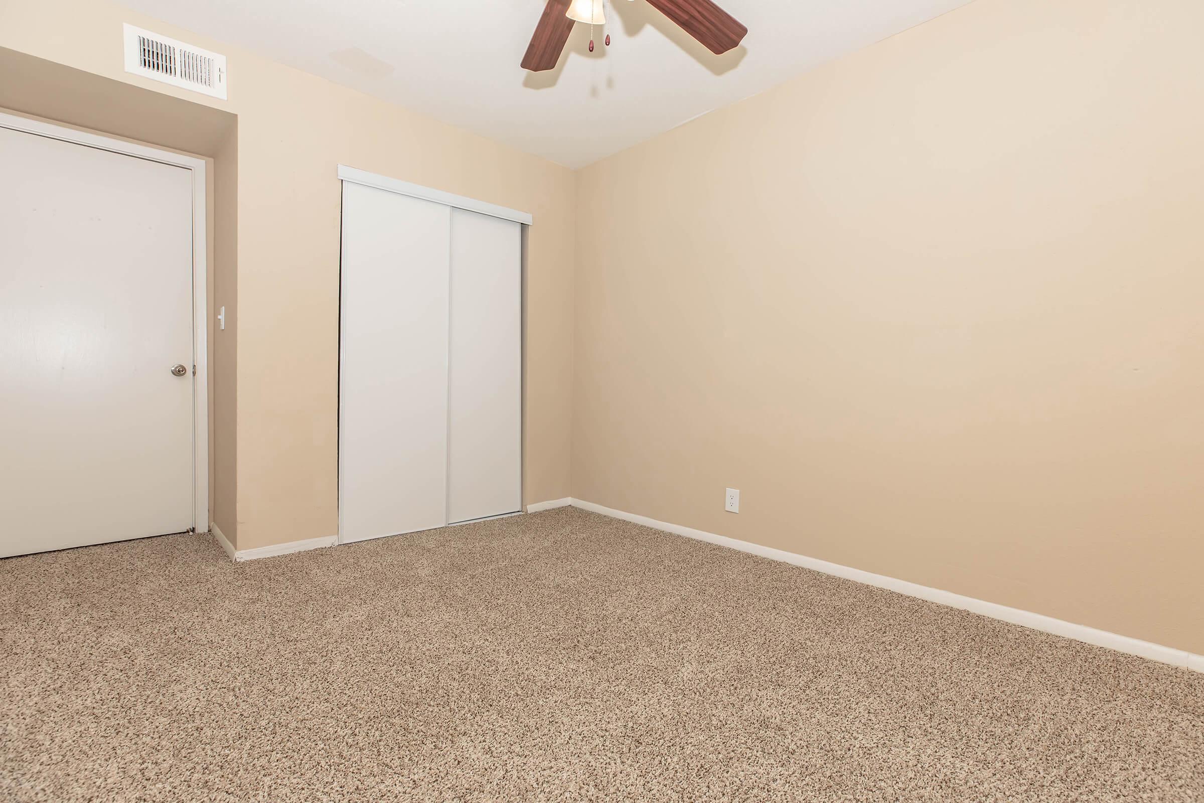 A vacant room with beige walls and carpet, featuring a ceiling fan and a closet with sliding doors. The room has a single door leading to another space and minimal decor, creating a clean, simple atmosphere.