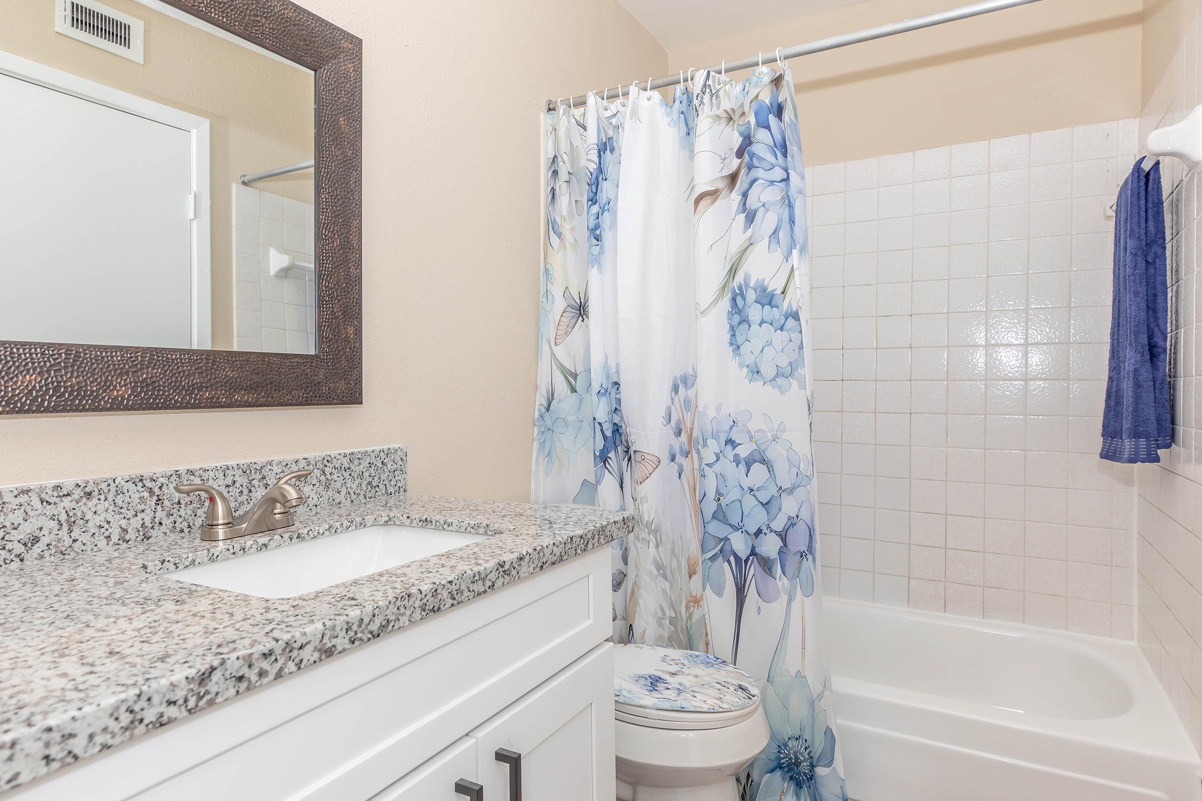 A clean bathroom featuring a white vanity with a granite countertop, a large mirror, and a floral shower curtain. The bathtub is white, and there's a blue towel hanging on the wall. The walls are a light neutral color, enhancing the overall brightness of the space.