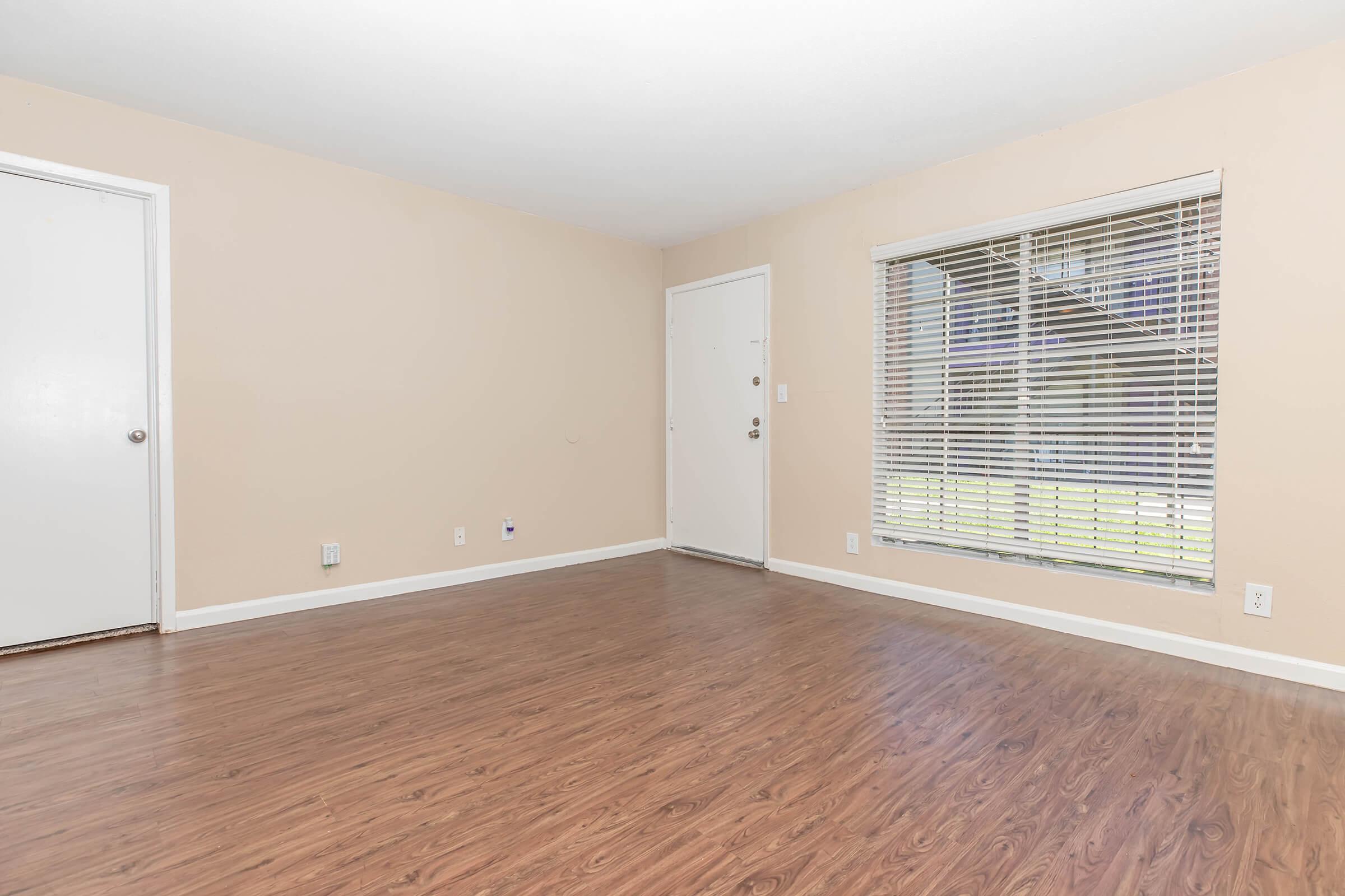 A vacant room with a light beige wall color and laminate wood flooring. There is a white door on the left and a window with blinds on the right, allowing natural light to enter. The overall space appears clean and ready for new furniture or decoration.