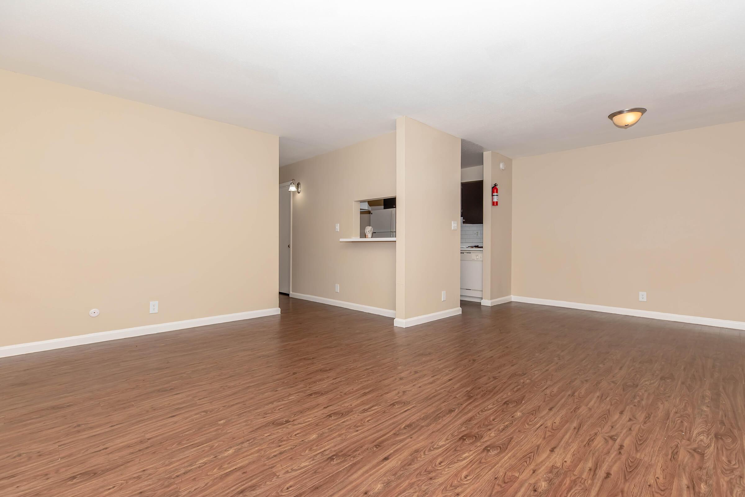 A spacious room with light beige walls and wooden flooring. The area is well-lit, featuring a ceiling light and an open layout. On one side, there is a bar-like counter that leads to a kitchen area, which is partially visible. The overall atmosphere is inviting and modern.
