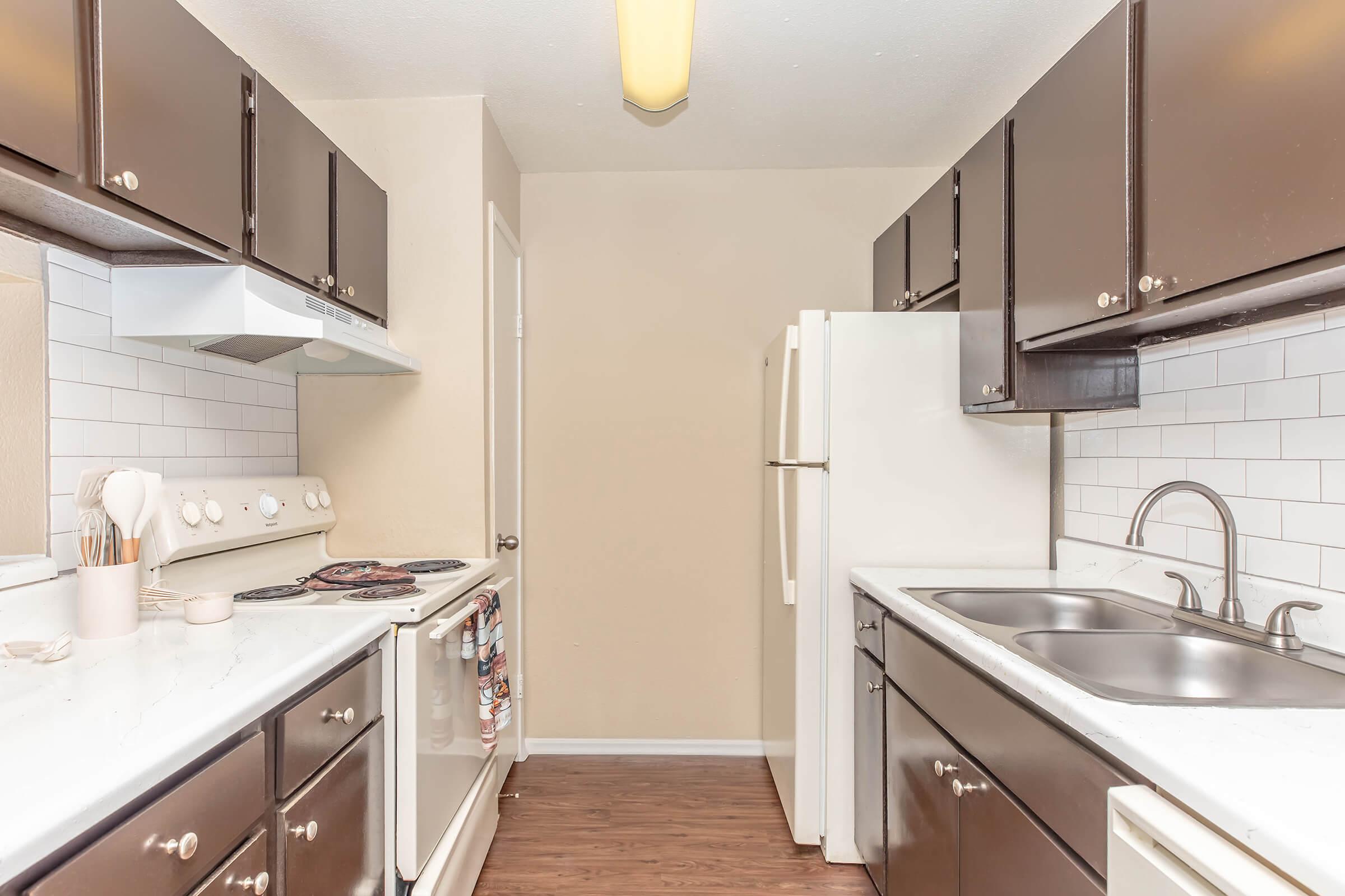 Modern kitchen featuring dark cabinetry, white tile backsplash, stainless steel sink, and a stove. Includes a refrigerator and ample counter space, with wood-style flooring. The walls are painted a light beige, creating a warm and inviting atmosphere.