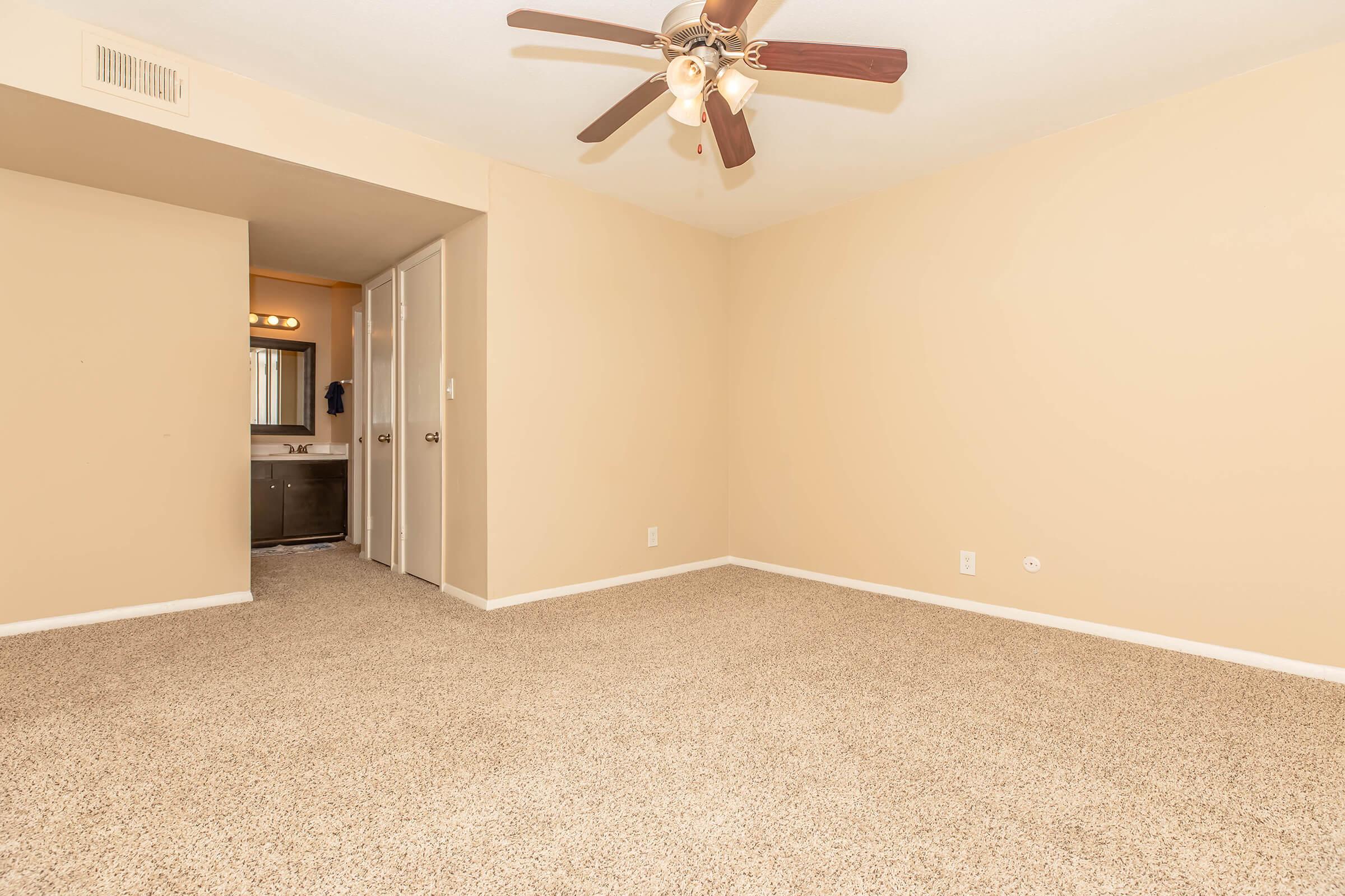 A spacious, empty room with light beige walls and carpet. A ceiling fan is visible. To the left, there is an open doorway leading to a bathroom area with lighting. The room is bright and uncluttered, providing a blank canvas for decoration and furnishing.