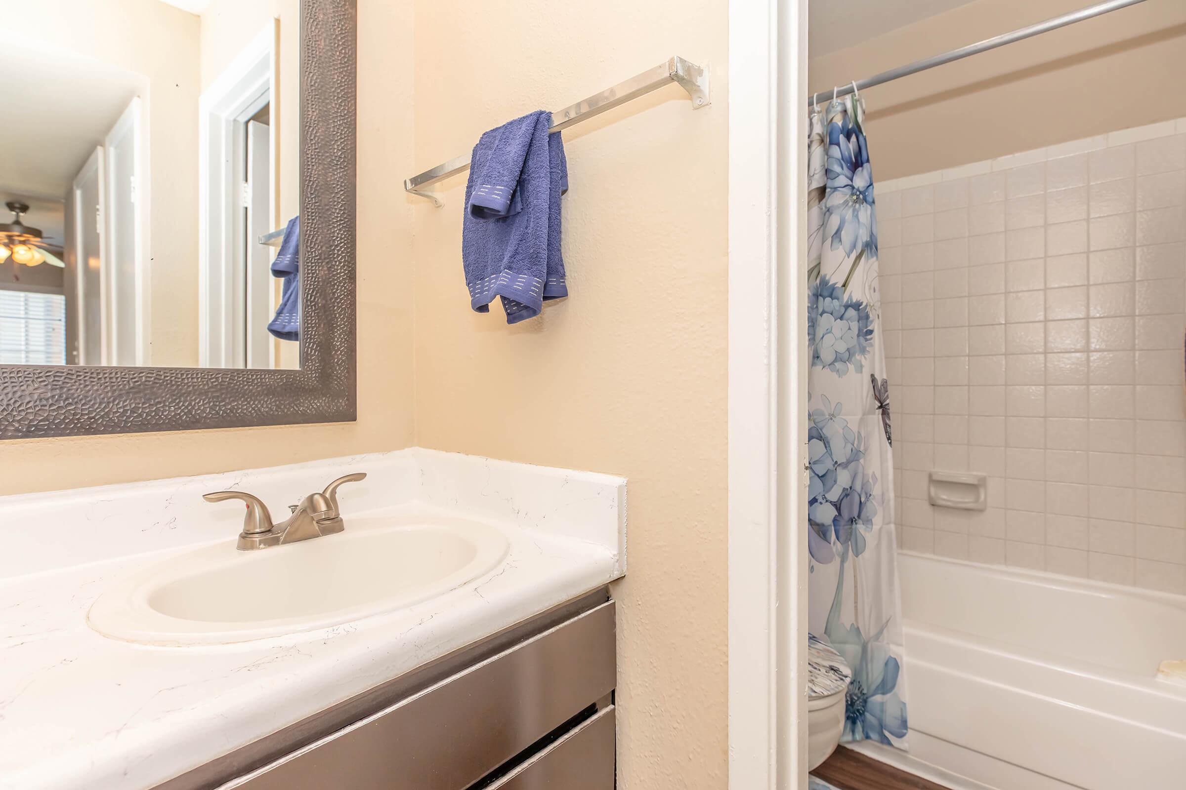 A well-lit bathroom featuring a sink with a faucet, a mirror with a decorative frame, and a towel rack with blue towels. In the background, a shower curtain with a floral design partially covers a bathtub. Light-colored walls and floors contribute to a fresh, clean ambiance.
