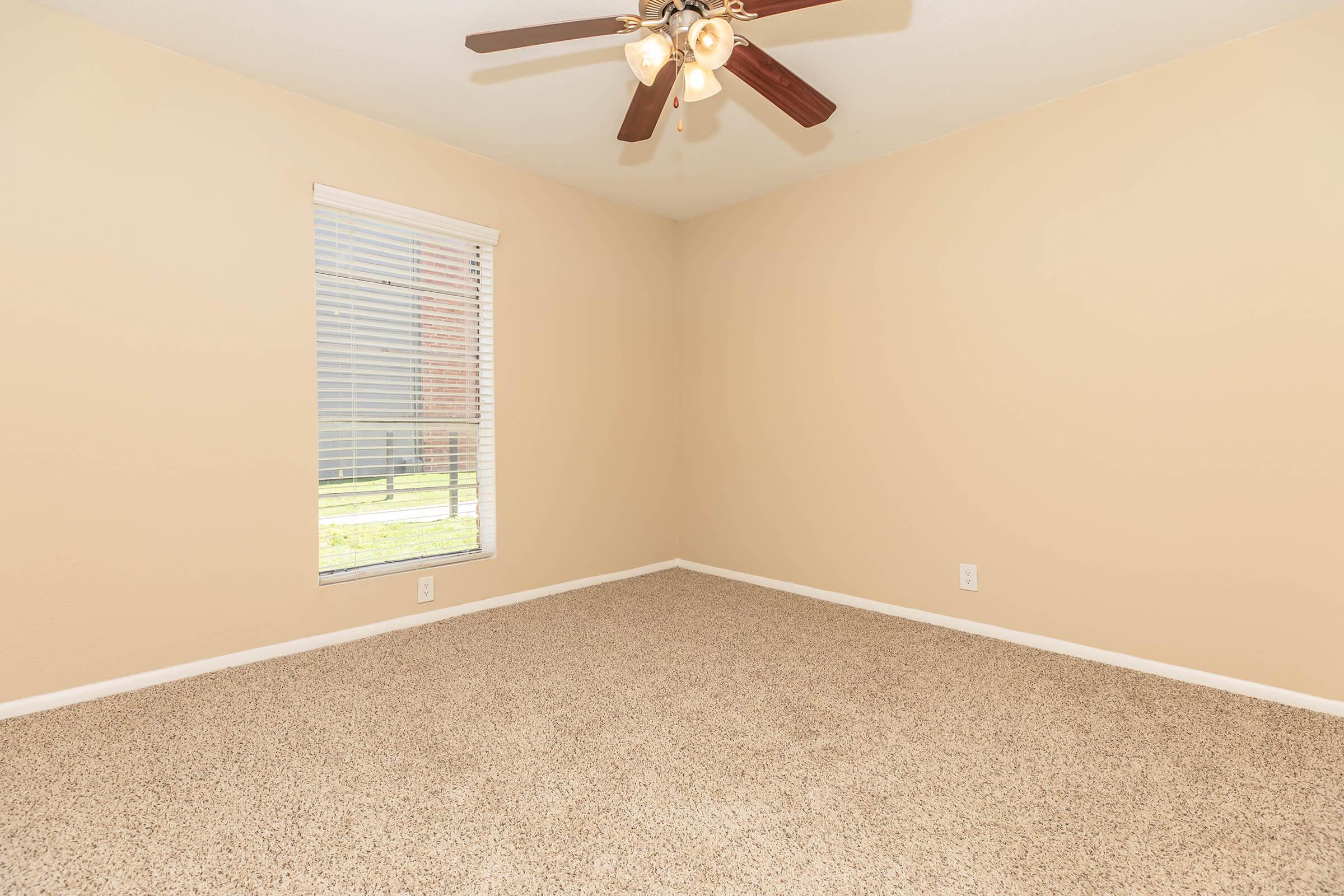 Empty room with beige walls and carpet, featuring a ceiling fan. A window with blinds allows natural light to enter, illuminating the space. The room has no furniture, creating an open and versatile atmosphere.