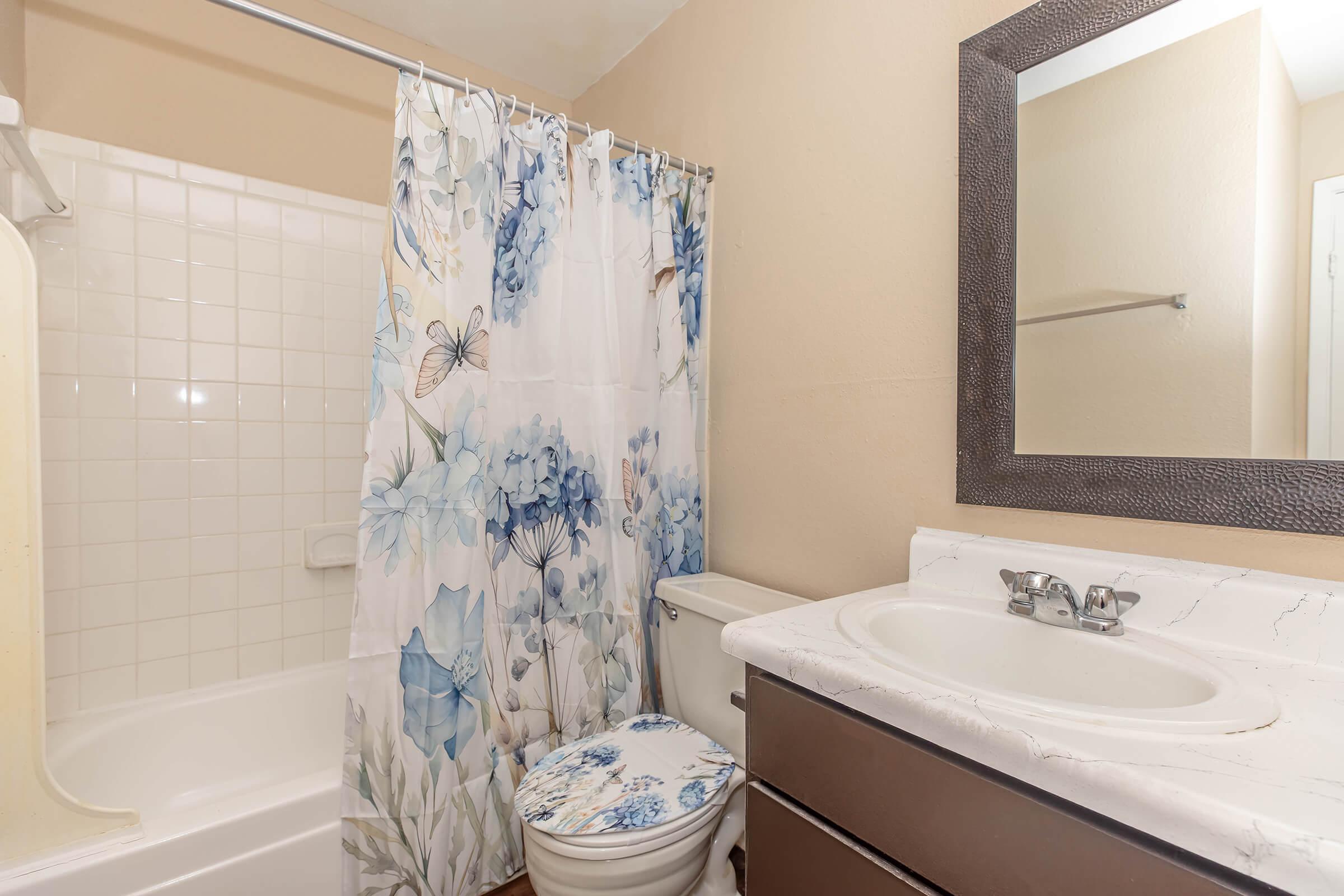 A clean, small bathroom featuring a bathtub with a shower, a floral-patterned shower curtain, a white toilet, and a vanity with a marble countertop and a mirror. The walls are painted a light beige color, and the floor is wooden.