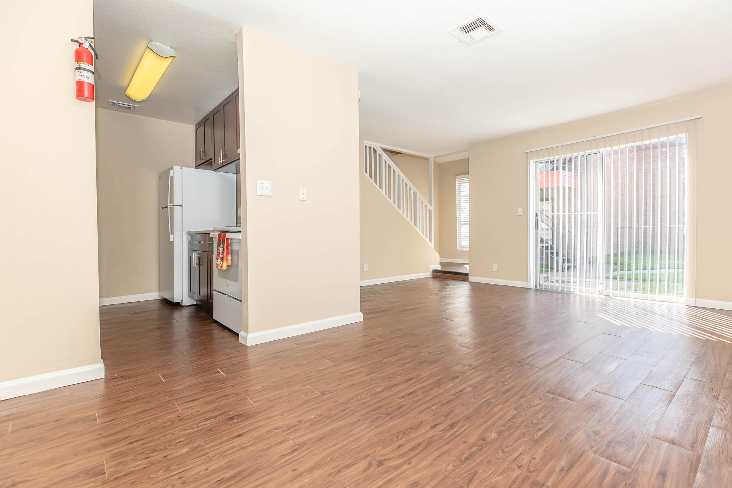 Interior view of a spacious living area featuring laminate flooring, a staircase, and access to a sliding glass door. The kitchen is visible with appliances like a refrigerator. Natural light comes in from the patio, creating a bright atmosphere.