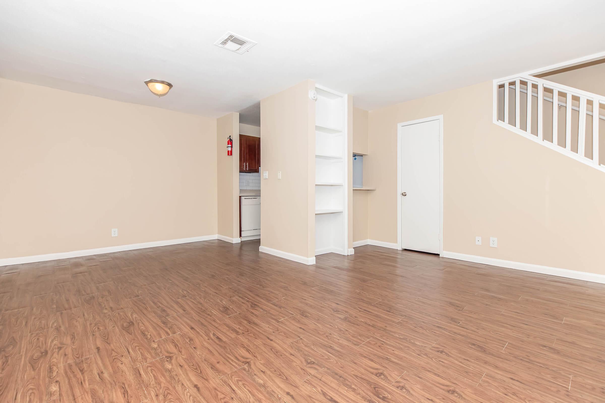 Interior view of a spacious, empty living area with light-colored walls and wooden flooring. A small staircase leads to an upper level on the right, and a doorway on the left opens to a kitchen area. There are built-in shelves along the wall, creating a clean and open space ready for furnishing.