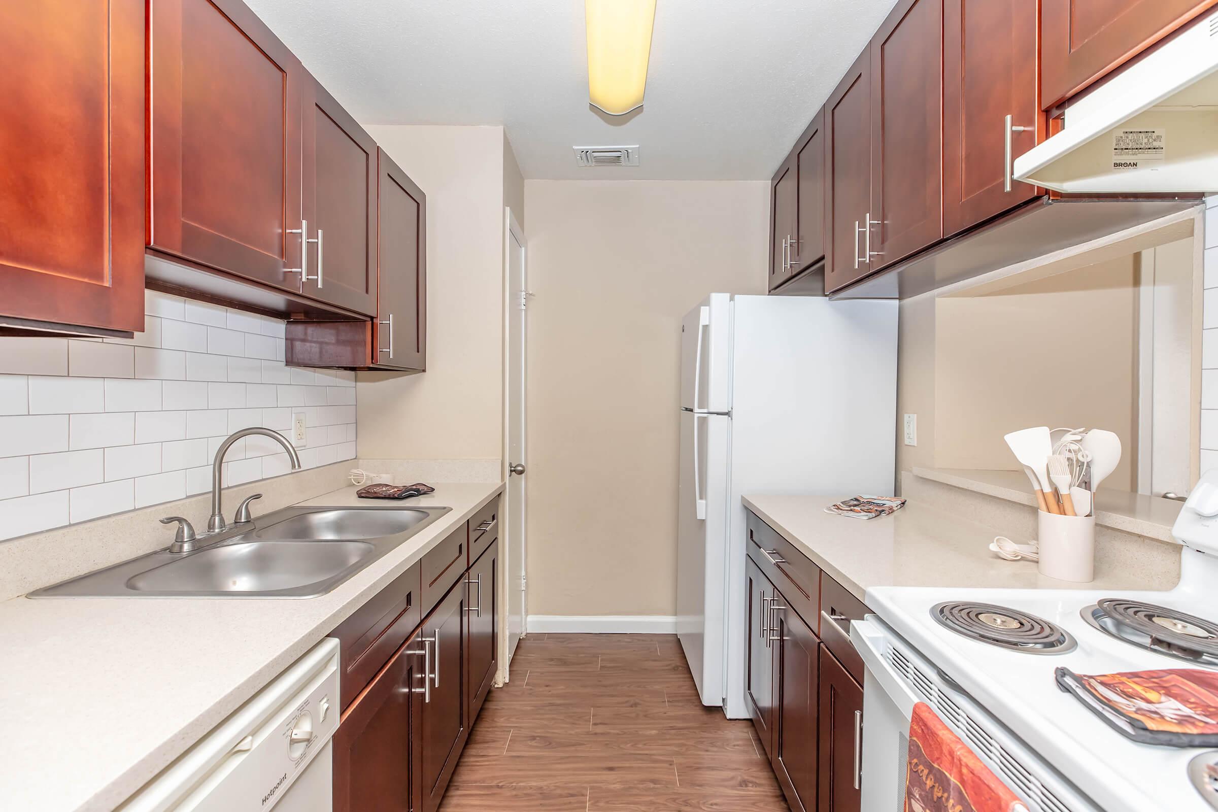 Interior view of a modern kitchen featuring dark wood cabinetry, a double sink, white appliances, and quartz countertops. The space includes a stove, oven, and refrigerator, with a neutral wall color and pendant lighting, creating a bright and functional cooking area.