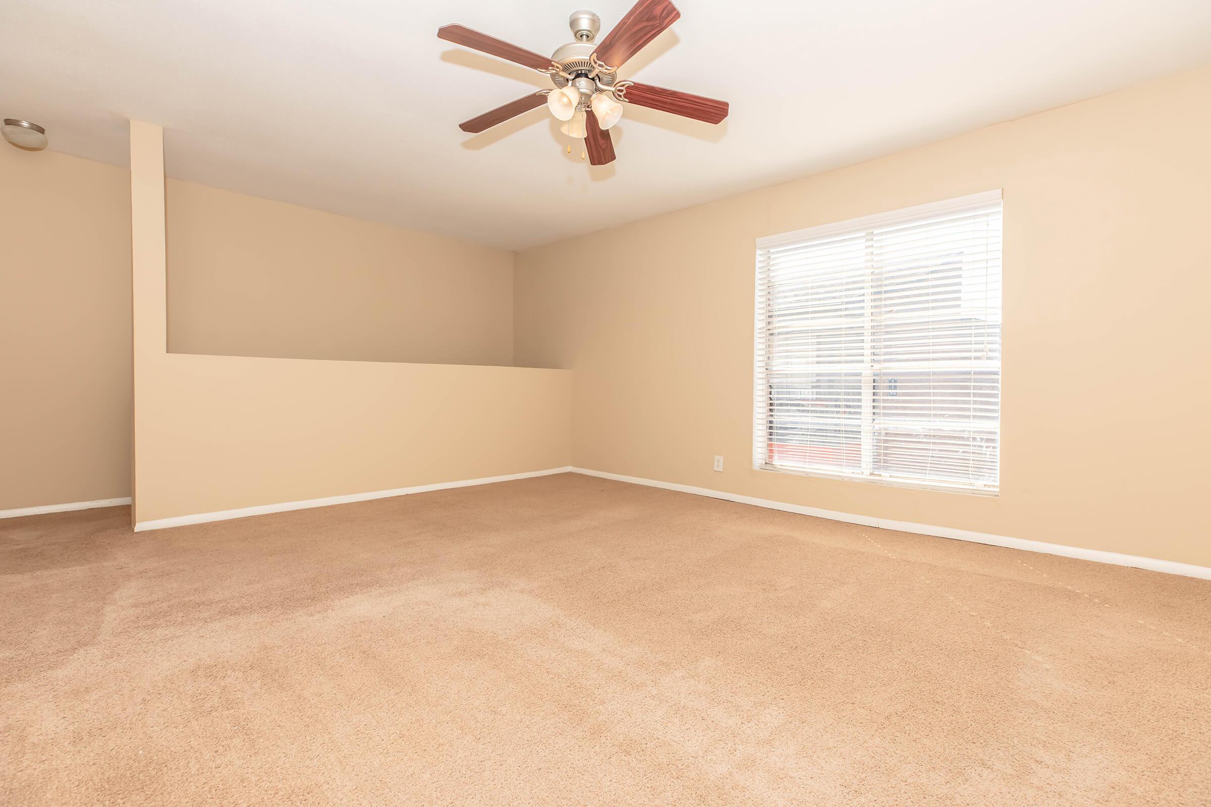 Spacious empty living room featuring light beige walls, a ceiling fan with four lights, and a large window with white blinds allowing natural light. The floor is covered with light brown carpet, creating a warm and inviting atmosphere.