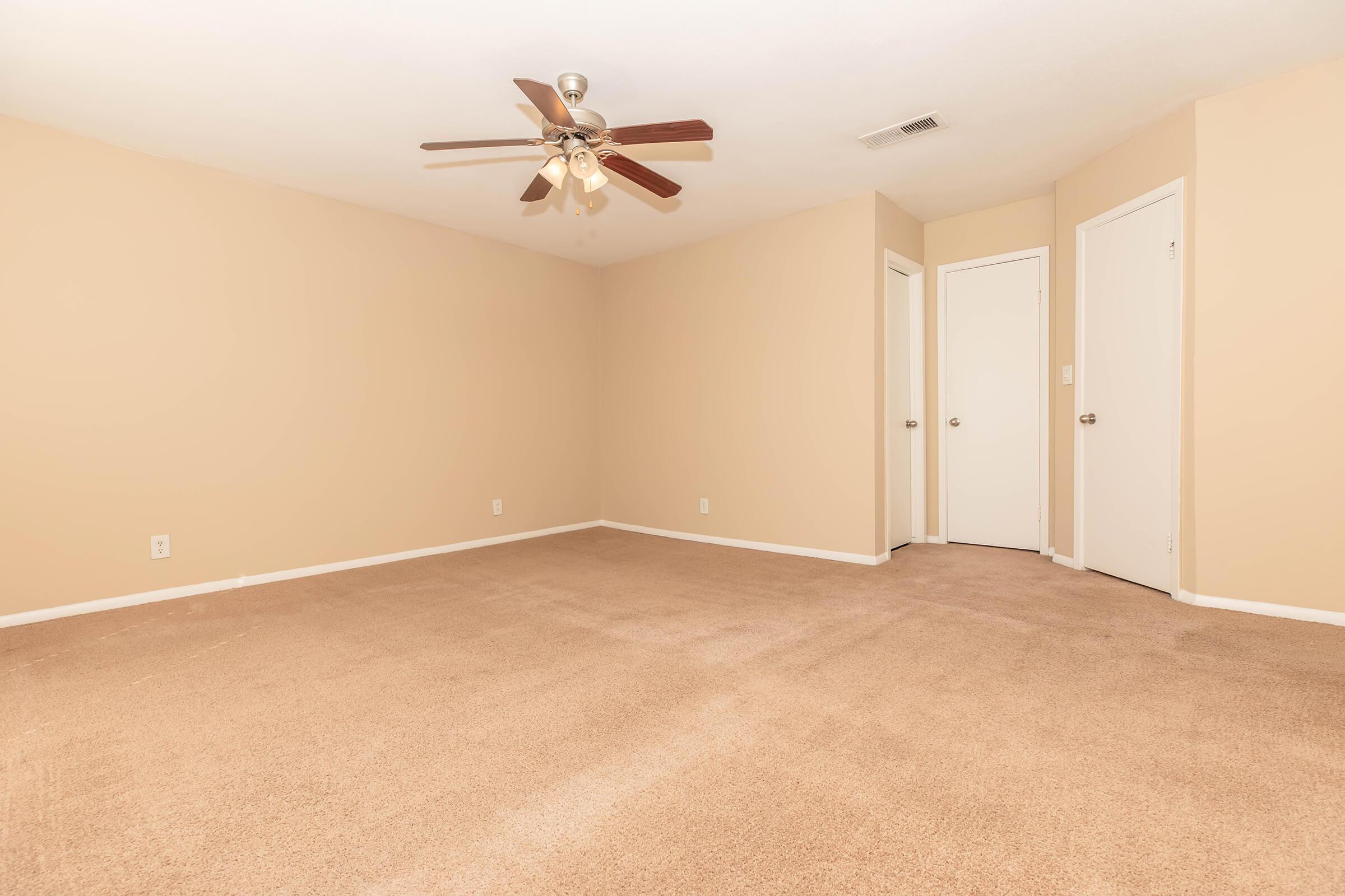 A spacious, empty room with beige walls and carpet. It features a ceiling fan with light fixtures, a corner with two closed doors, and ample natural light, creating an airy atmosphere. Ideal for various uses, such as a bedroom or living space.