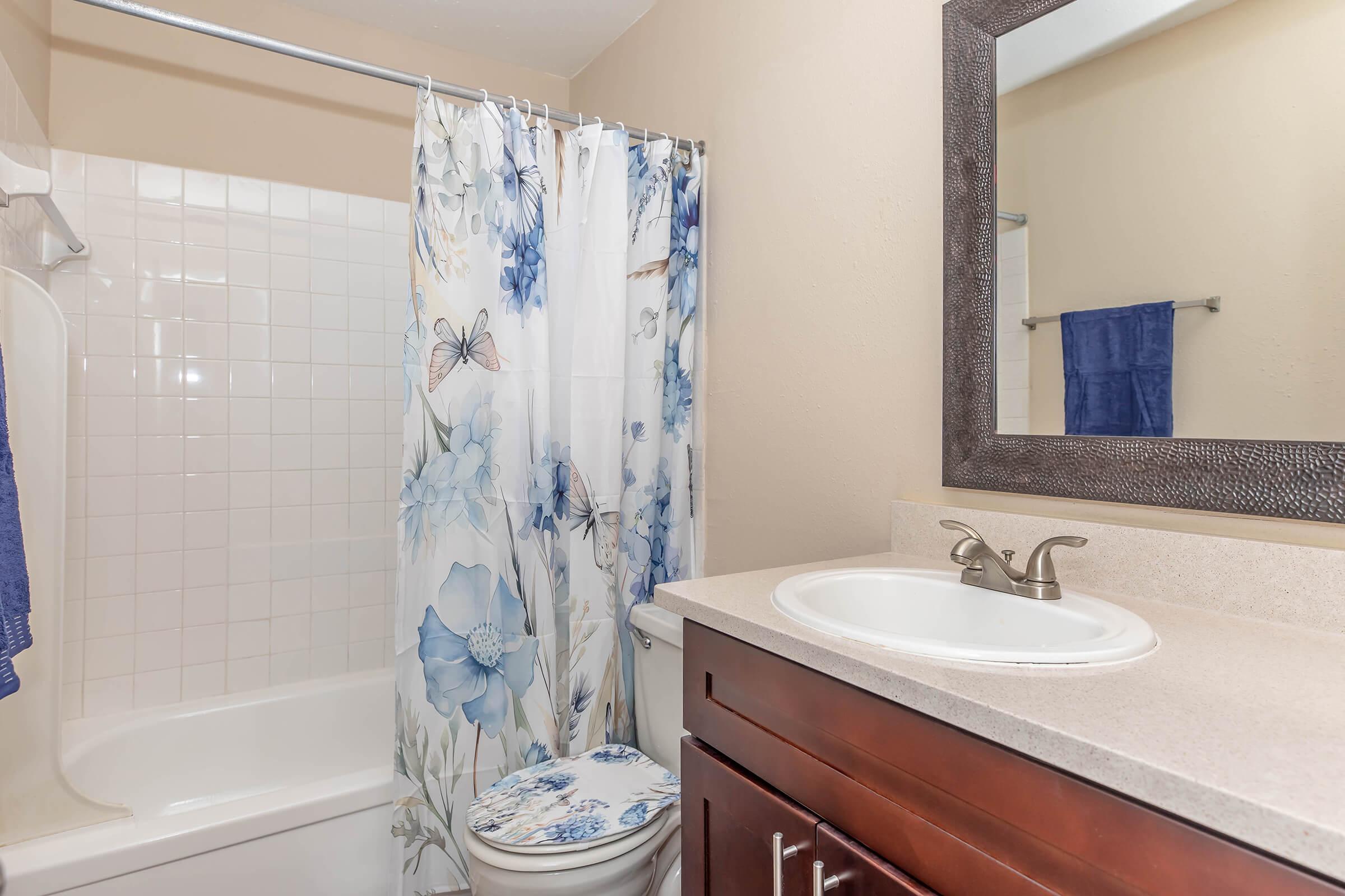 A clean bathroom featuring a white bathtub with a floral shower curtain, a sink with a modern faucet, and a large mirror above the sink. The countertop is light-colored, and there's a blue towel hanging on the wall, adding a touch of color to the space.