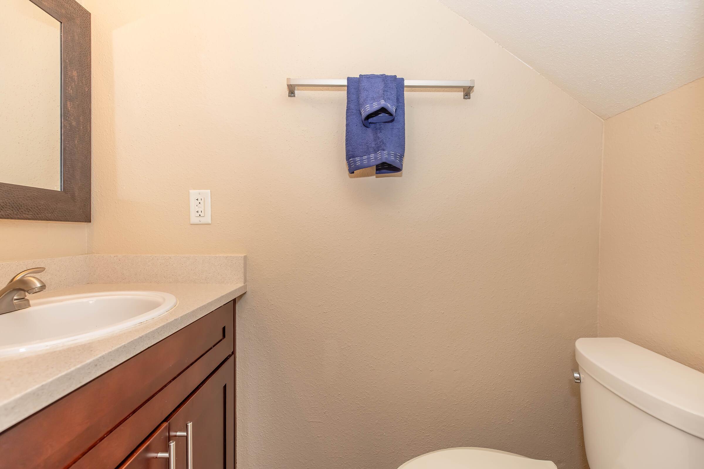 A small bathroom featuring a light-colored wall, a mirror above a sink with a countertop, and a dark wooden vanity. A silver towel rack holds a blue towel. A white toilet is positioned to one side, completing the simple, functional layout.