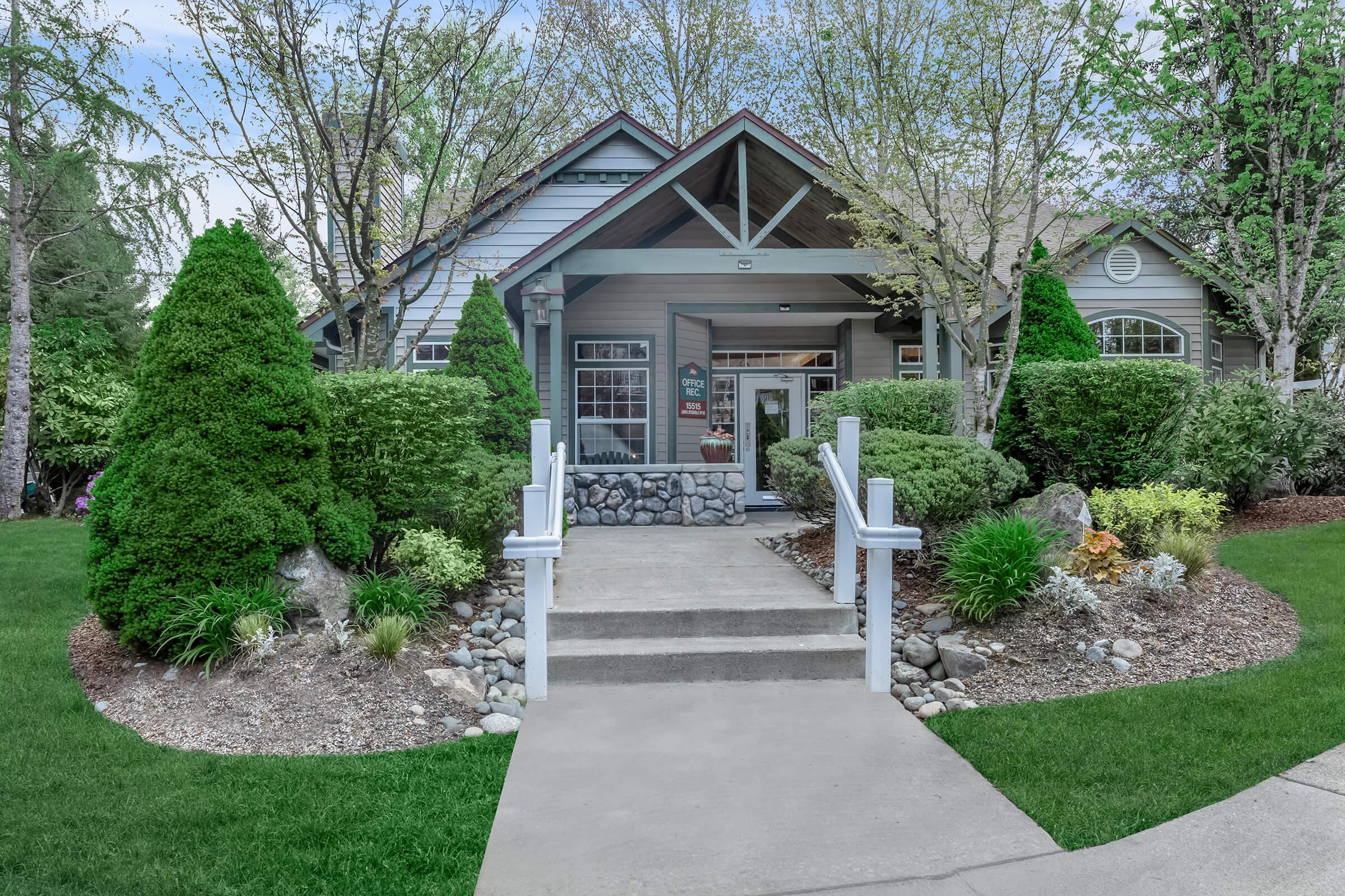 A welcoming front entrance of a home, featuring a stone pathway leading to a porch. The yard is well-manicured with neatly trimmed bushes and vibrant flower beds, surrounded by lush greenery and trees. The house has a blend of rustic and modern architectural elements with a pitched roof and large windows.