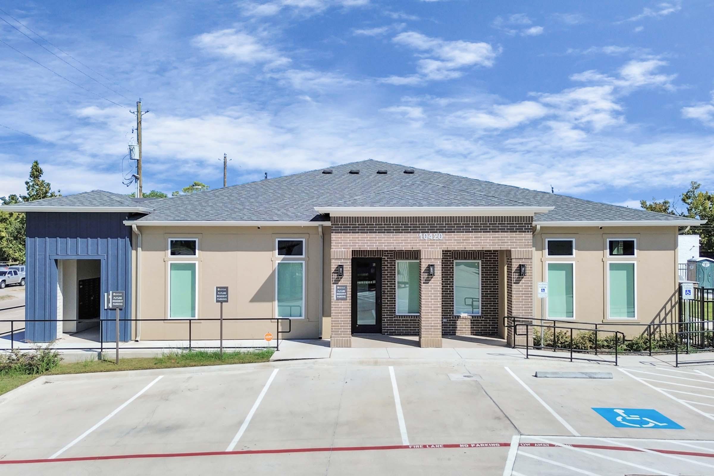 A modern single-story building with a smooth beige facade, brick accents, and a dark blue section to the left. It features large windows and a well-maintained parking area with accessible spaces. The sky is blue with a few scattered clouds, indicating a clear day.